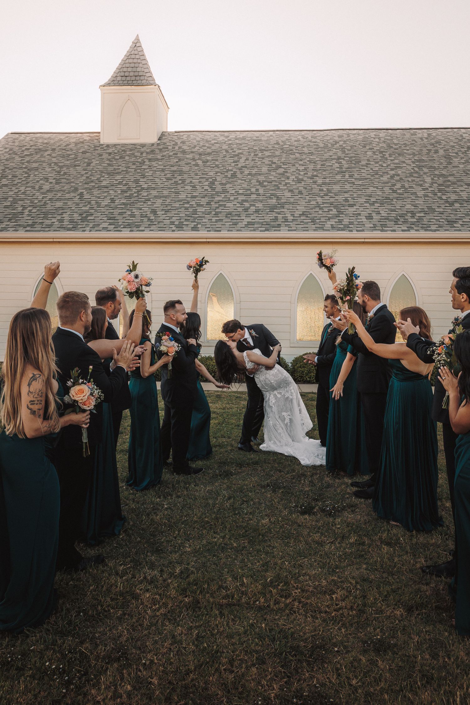 Wedding party celebrates as bride and groom share a romantic dip kiss outside a white chapel.