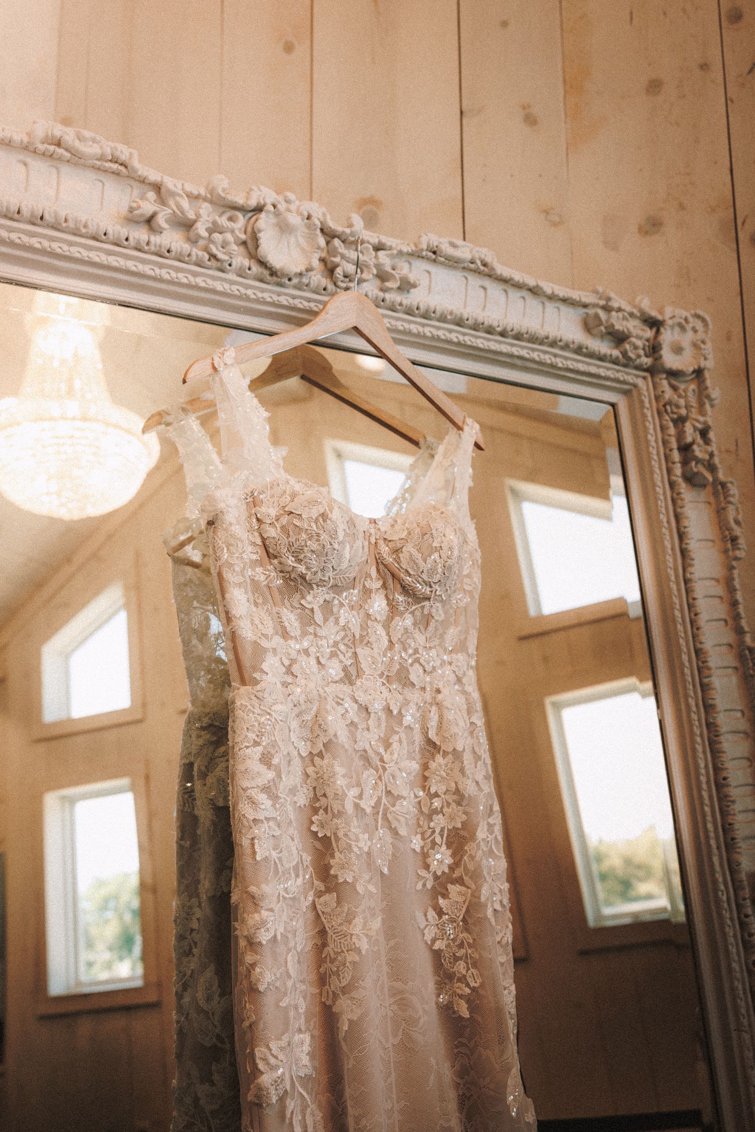 Intricate lace wedding gown hanging on a wooden hanger reflected in an ornate vintage mirror.