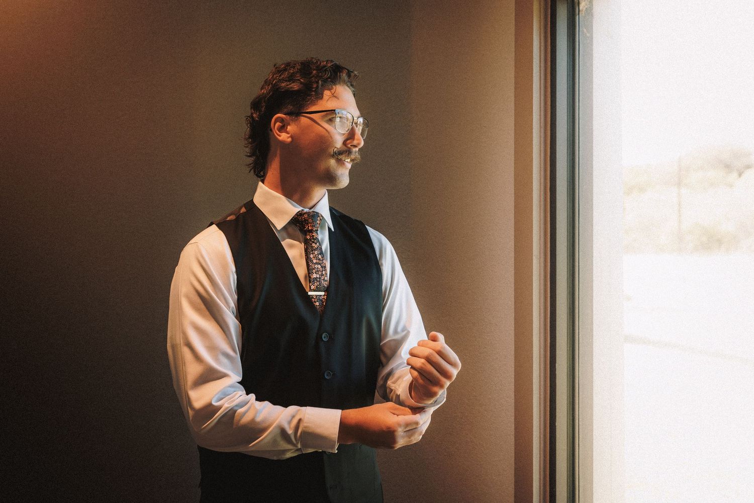 Dapper groom in vest and tie adjusting cufflinks while gazing out a window.