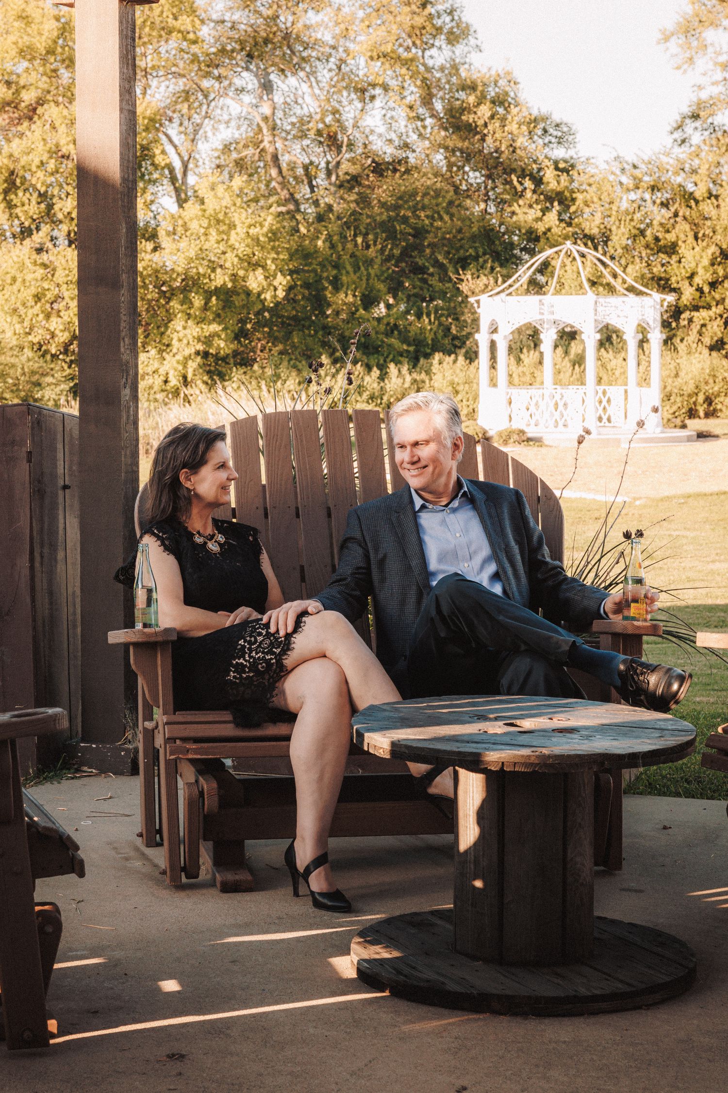 Couple relaxes on wooden chairs near a gazebo at a wedding reception venue.