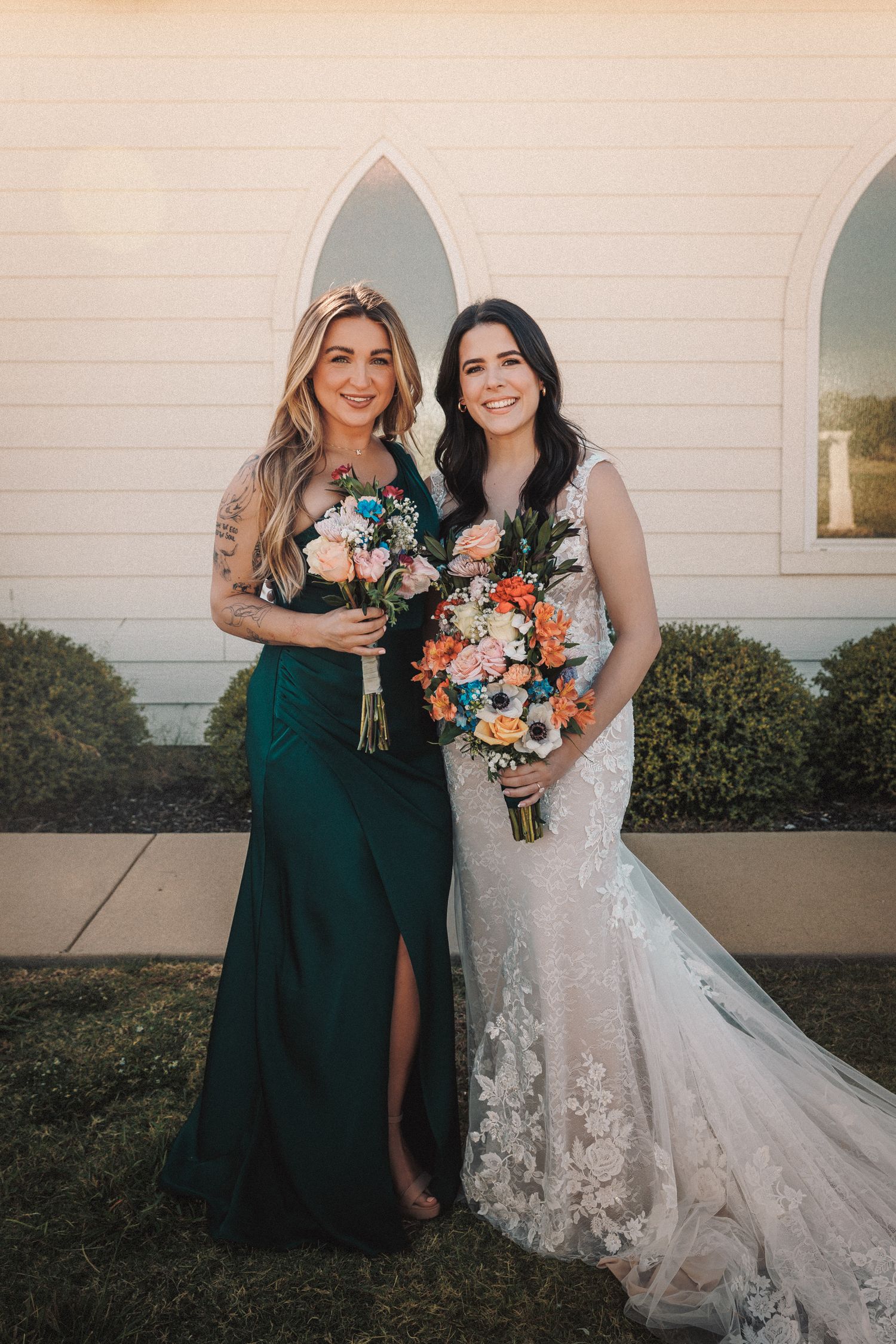 Bride in lace wedding gown posing with bridesmaid in dark green dress outside white church.