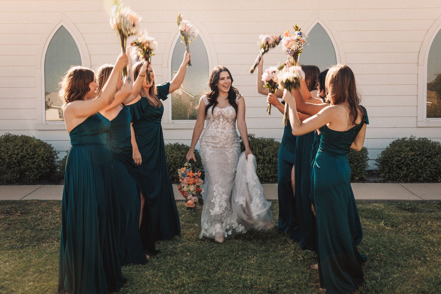Bride in lace wedding gown surrounded by bridesmaids in teal dresses raising bouquets in front of a white chapel.