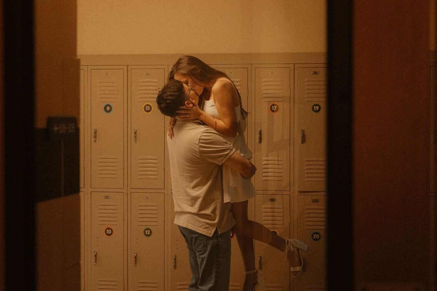 Couple sharing a romantic kiss in a warmly lit school hallway with beige lockers in the background.