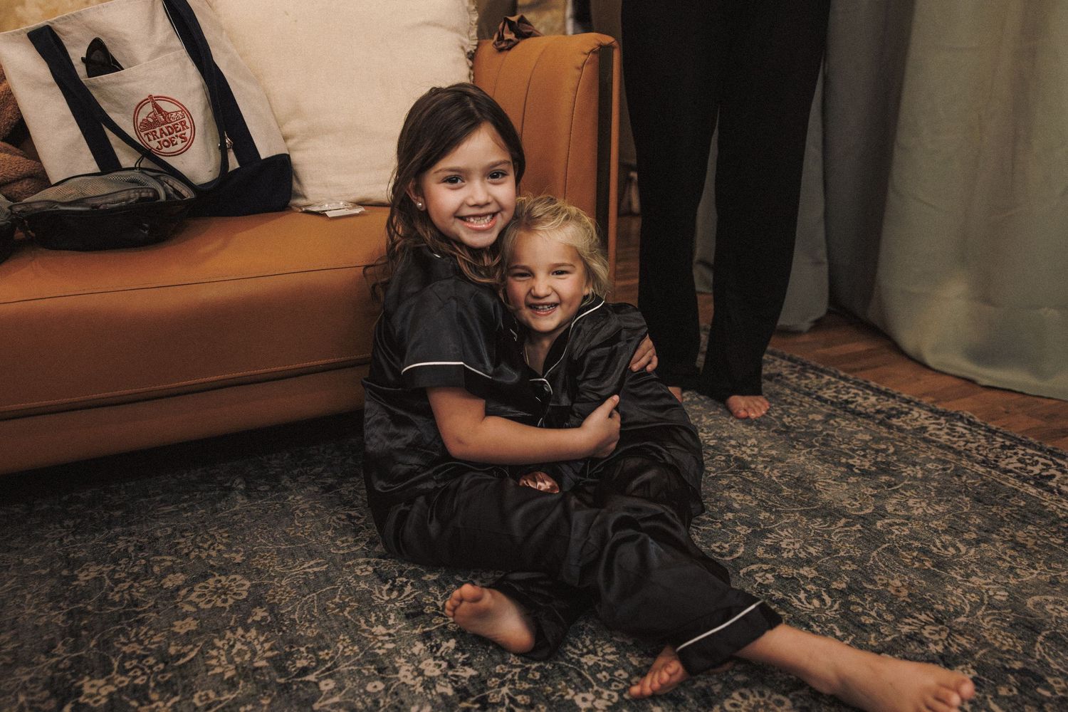 Two young girls in matching black pajamas sit on a patterned rug, smiling and embracing.