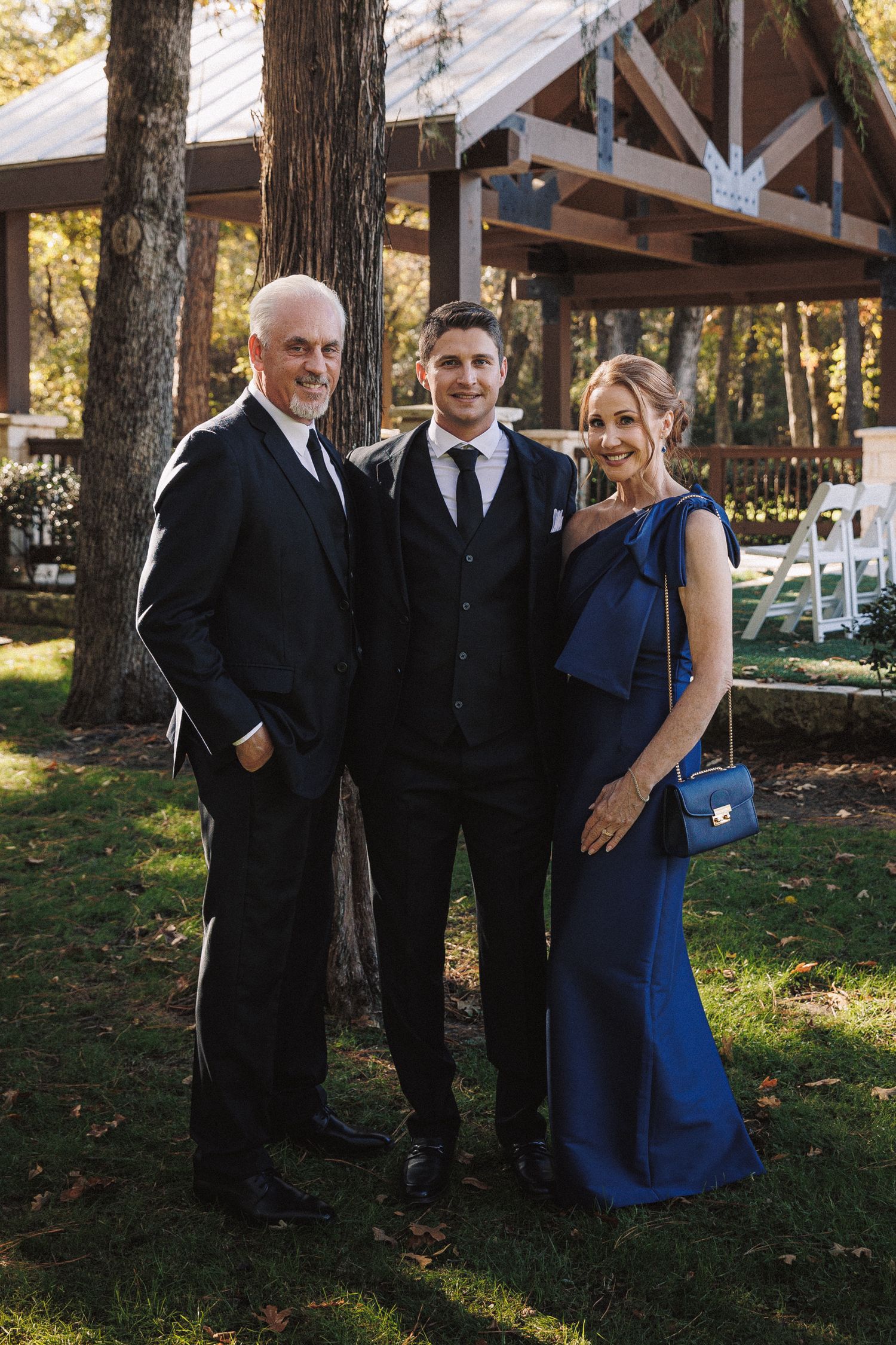 Groom in black suit poses with parents outdoors near a wooden pavilion on wedding day.