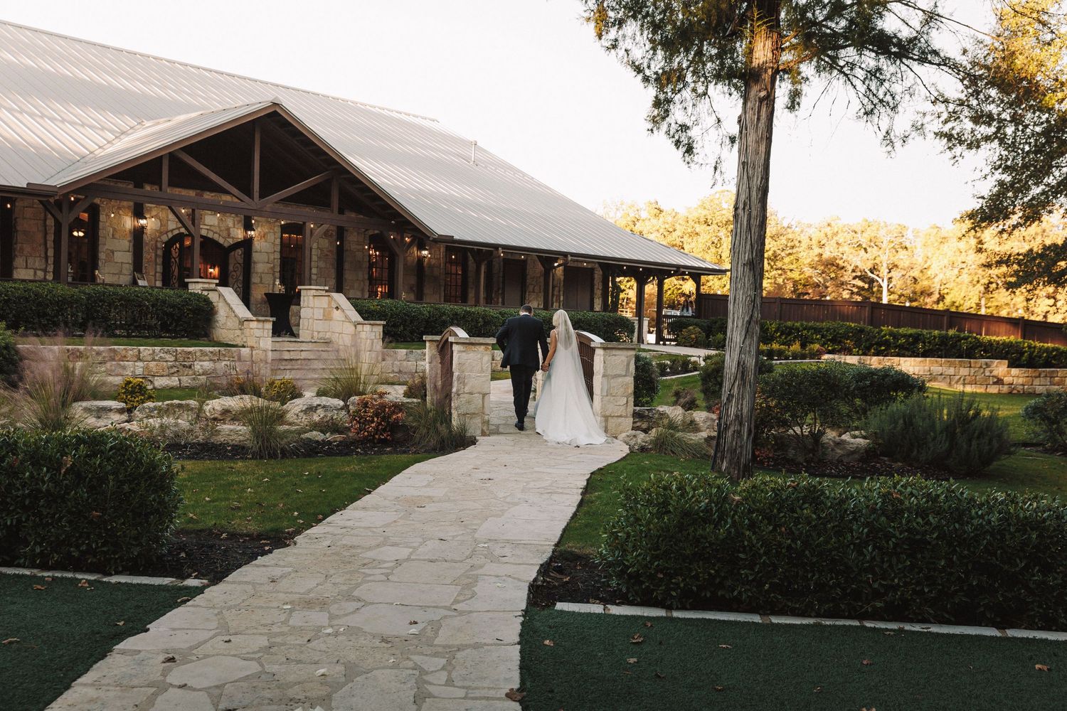 Bride and groom walking hand in hand toward rustic stone wedding venue at dusk.