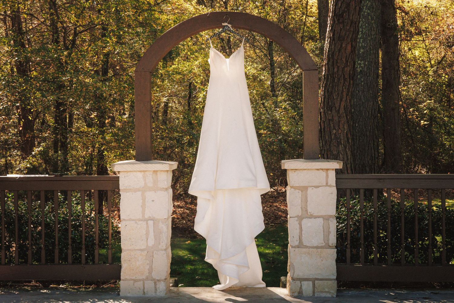 White wedding dress hanging on a stone arch surrounded by autumn trees in an outdoor garden setting.
