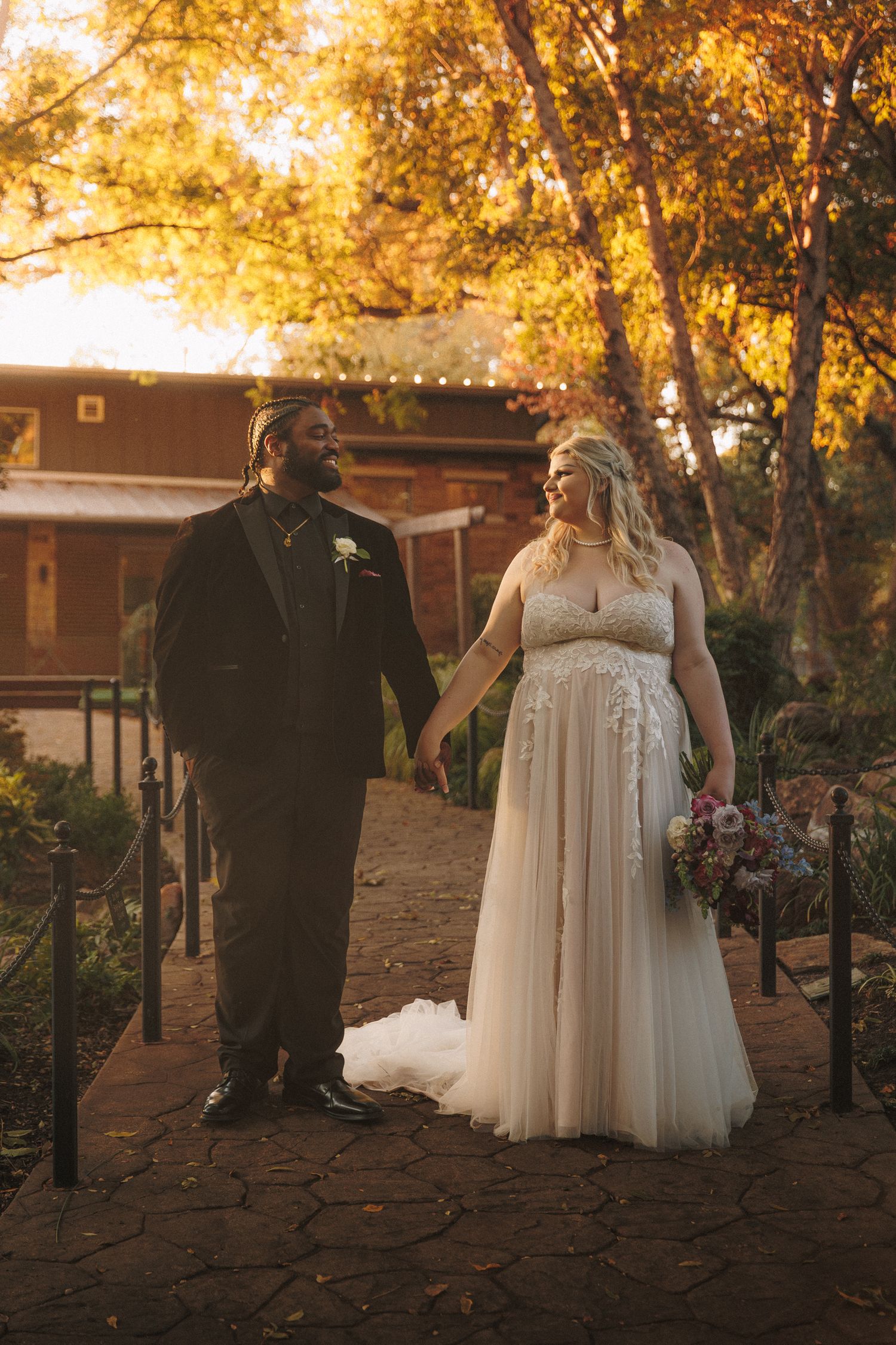 Interracial couple holding hands on garden path in golden autumn light on wedding day.