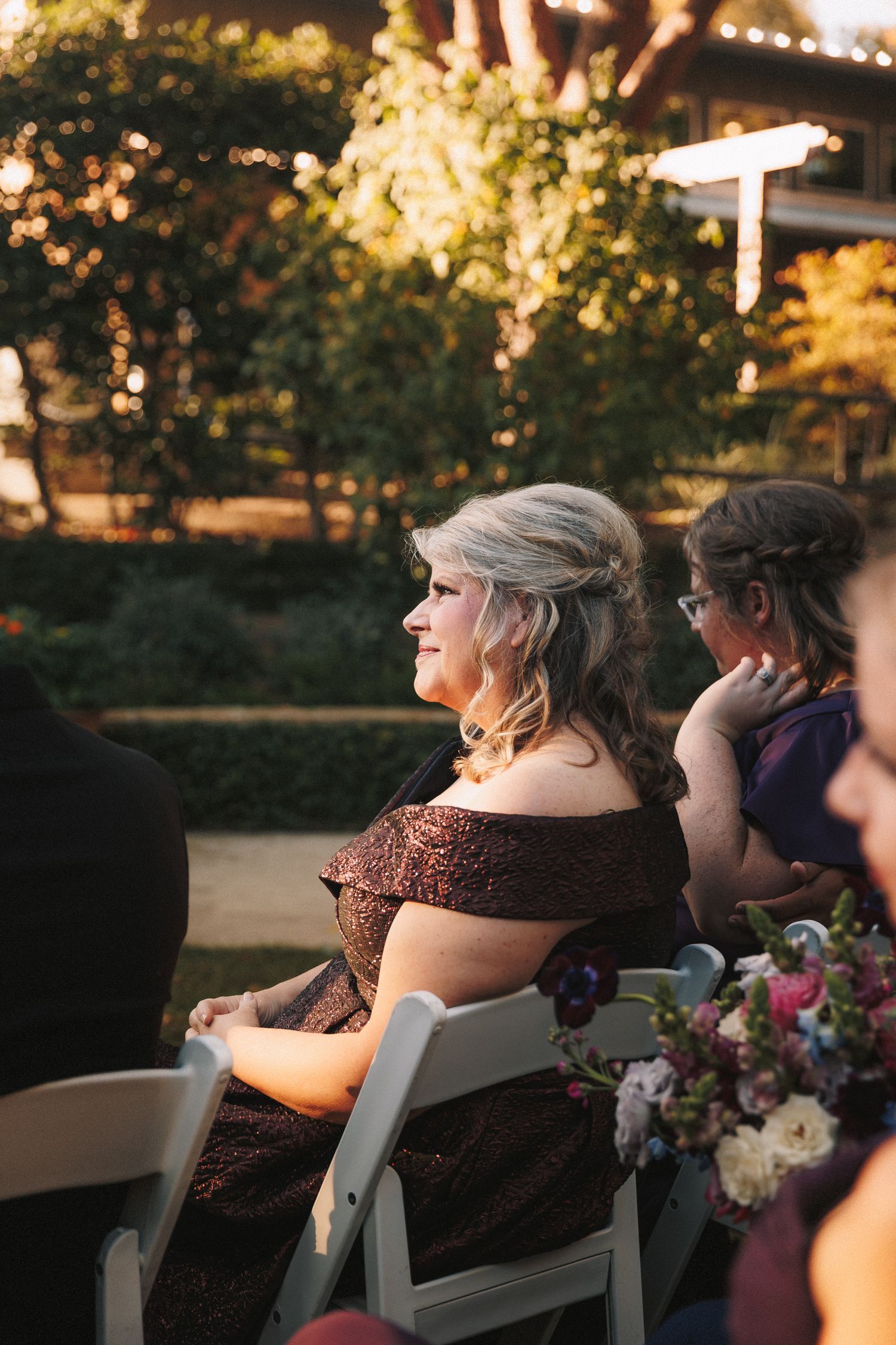 Elegant wedding guest in off-shoulder dark gown smiling during outdoor ceremony in golden afternoon light.