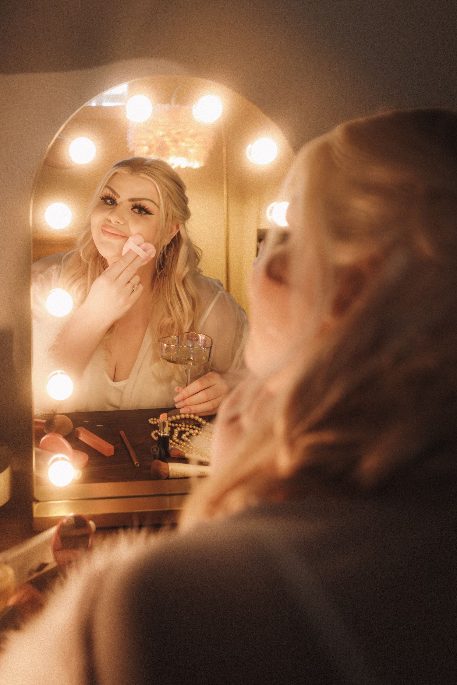 Bride applying makeup at a Hollywood vanity mirror with warm glowing bulbs holding champagne.