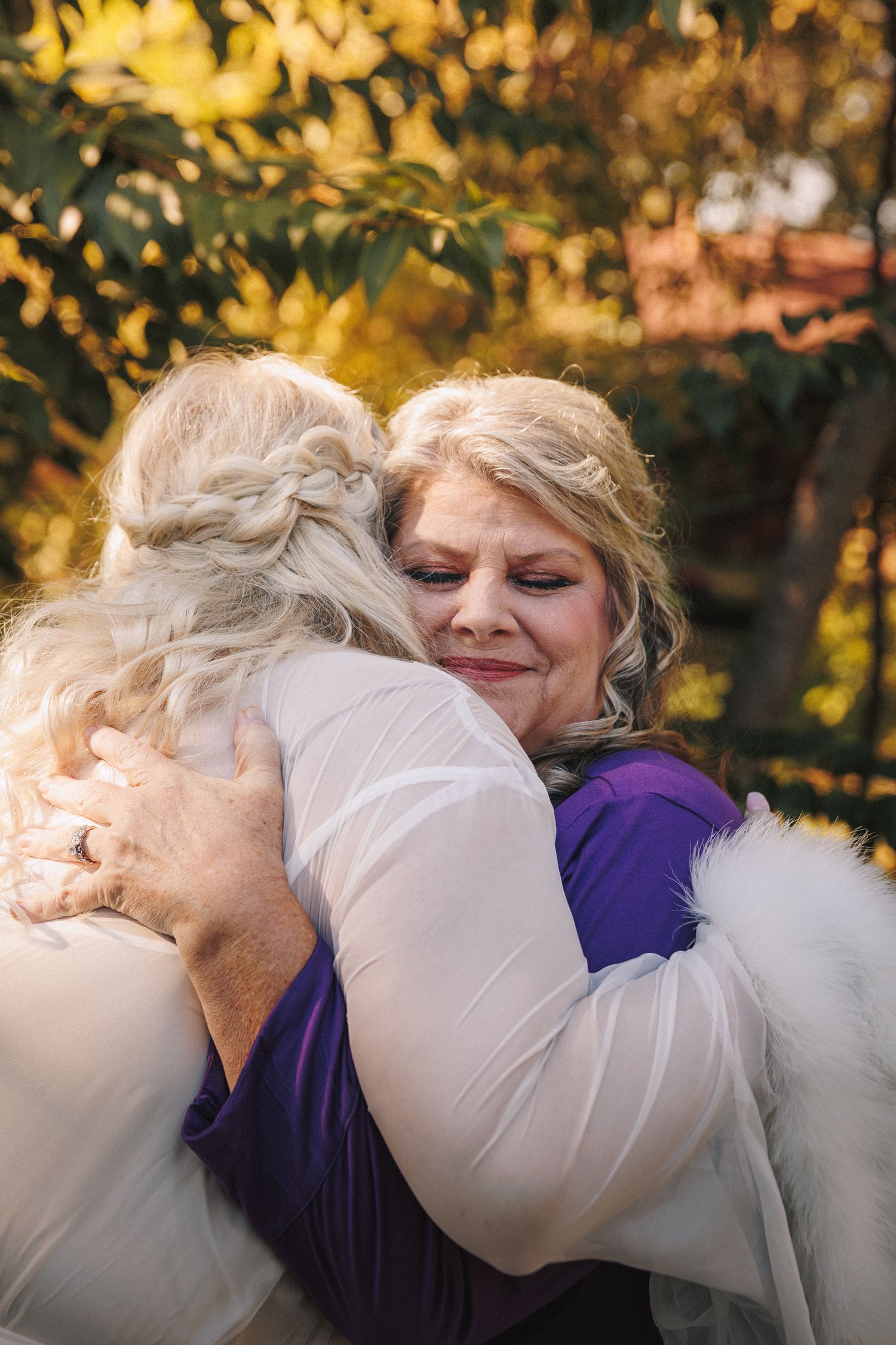 Emotional mother and bride sharing a heartfelt hug outdoors surrounded by golden autumn foliage.
