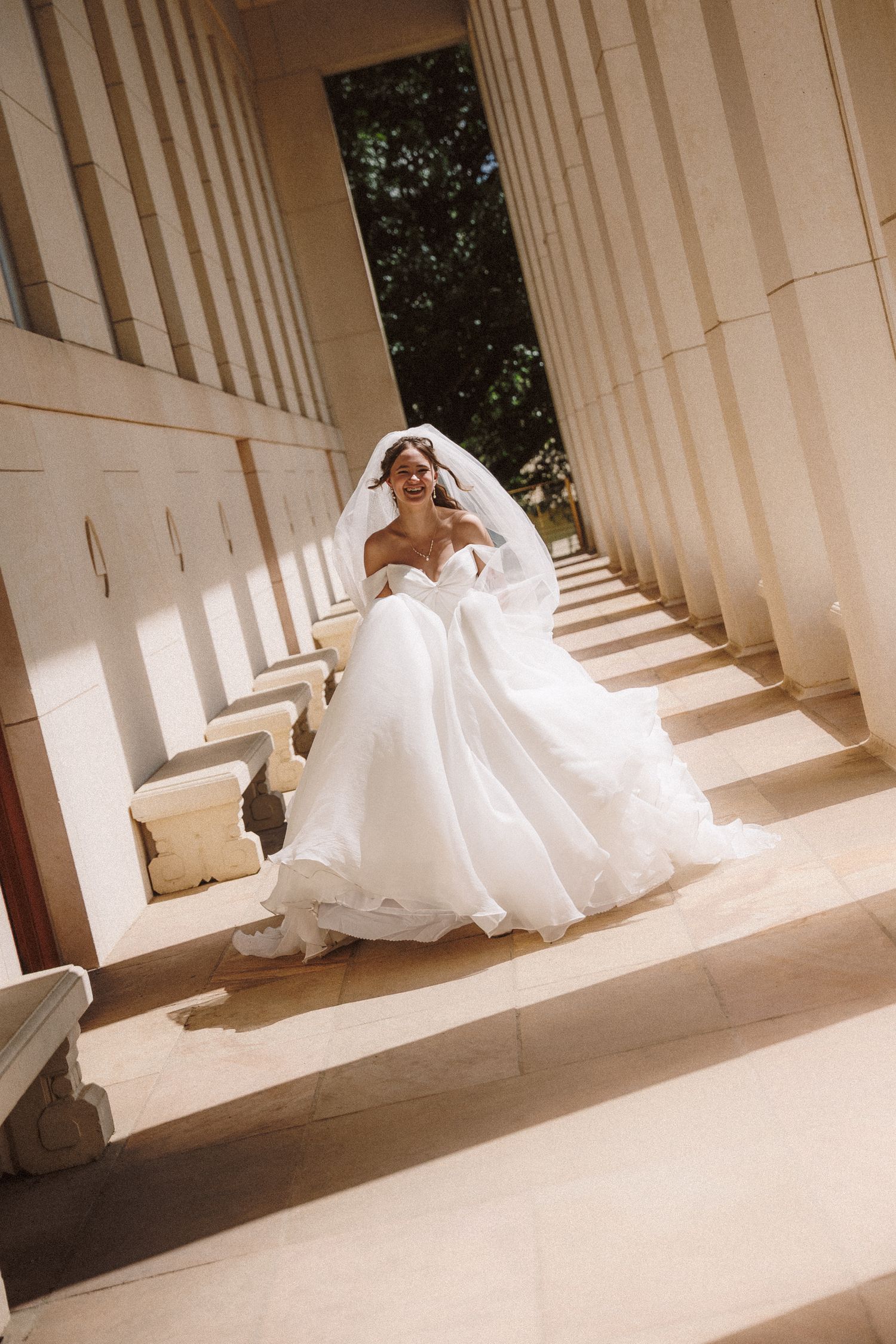 Bride in flowing white ball gown and veil walking between tall stone columns outdoors.