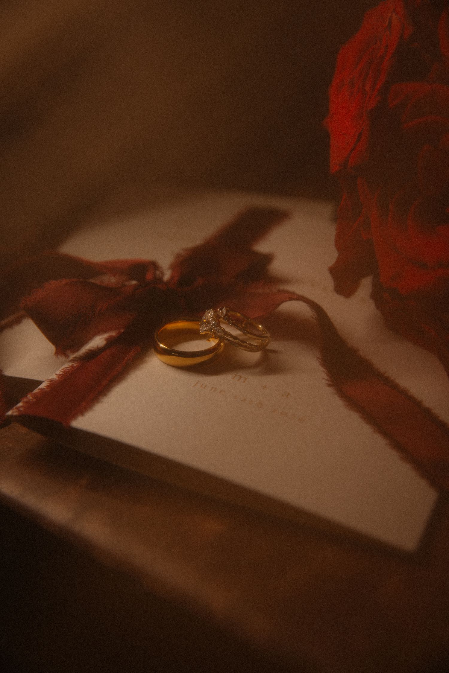 Two gold wedding rings resting on a white gift box tied with a red ribbon, with a rose in the background.