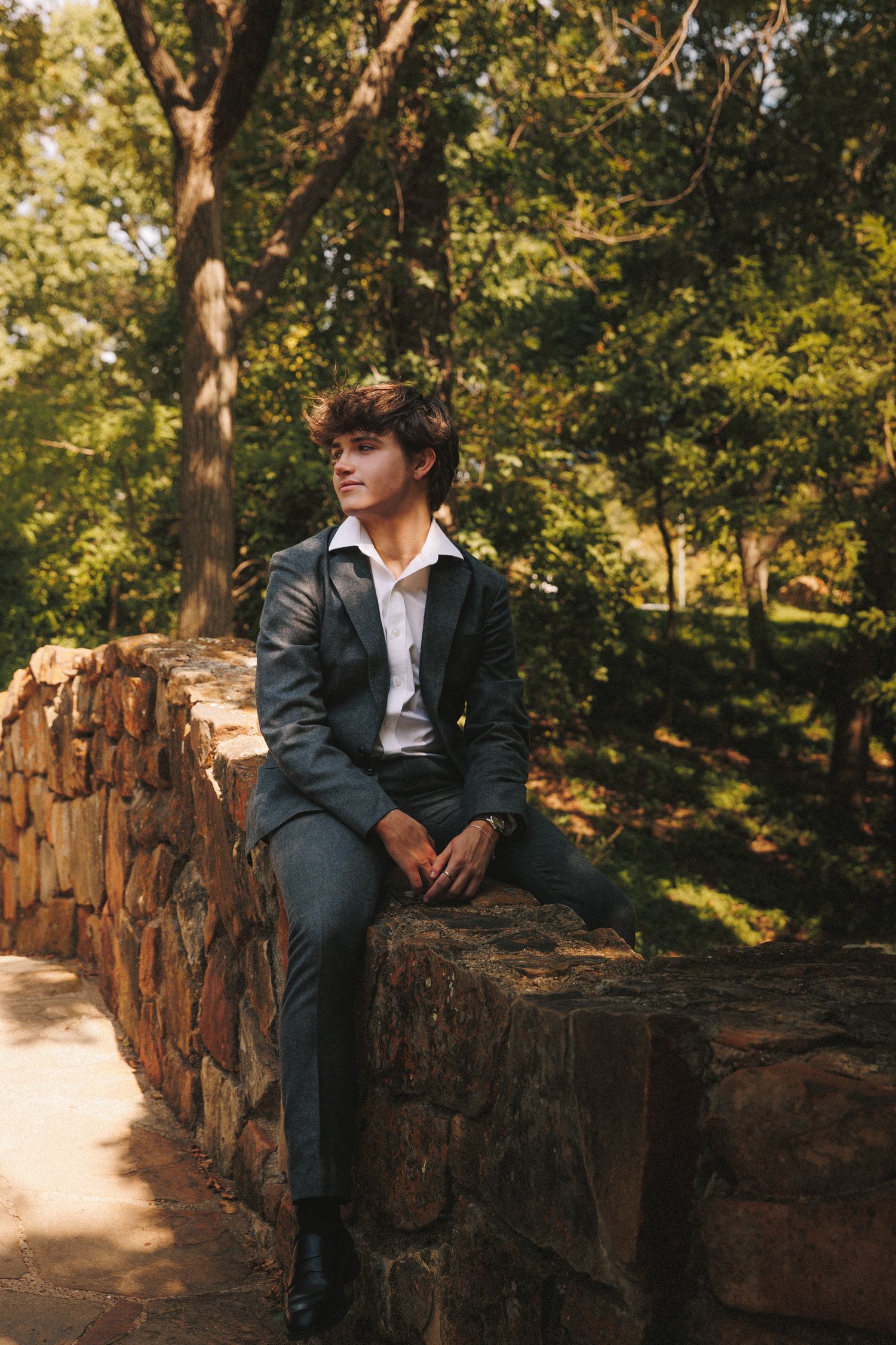A figure sits on a rustic stone wall surrounded by autumn trees in a park-like setting during golden hour.