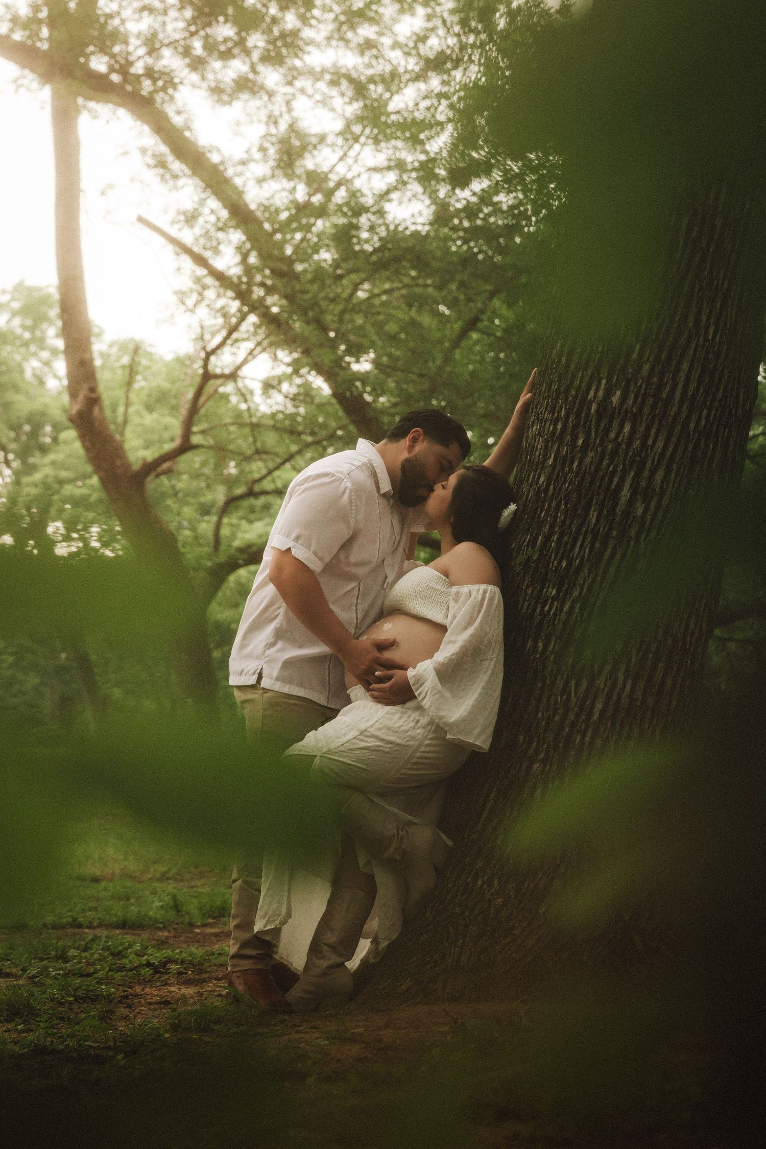 A romantic couple in white outfits shares an intimate moment against a tree trunk in a dreamy forest setting.