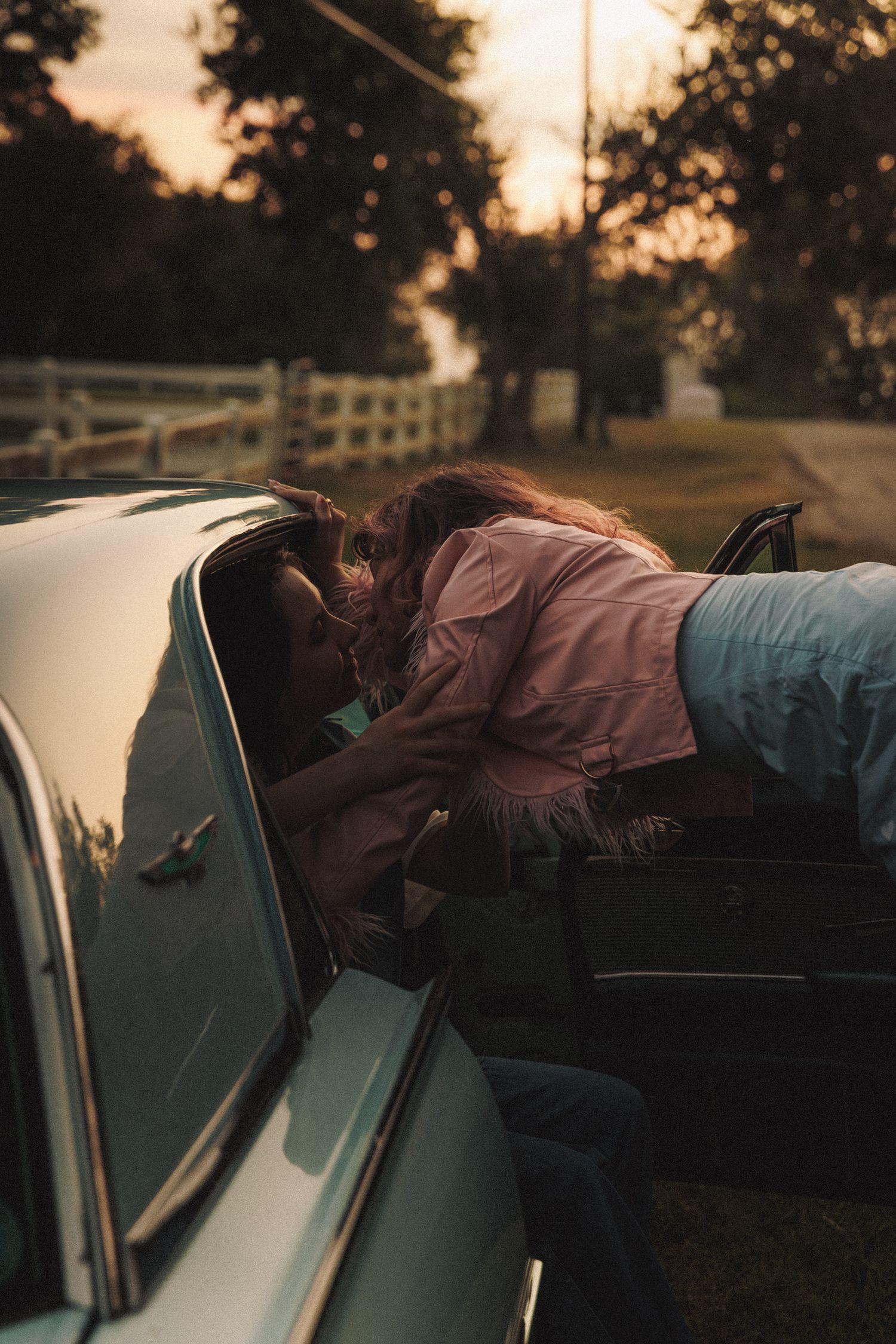 Someone leans through a car window at sunset in a rural setting with white fencing visible in the background.