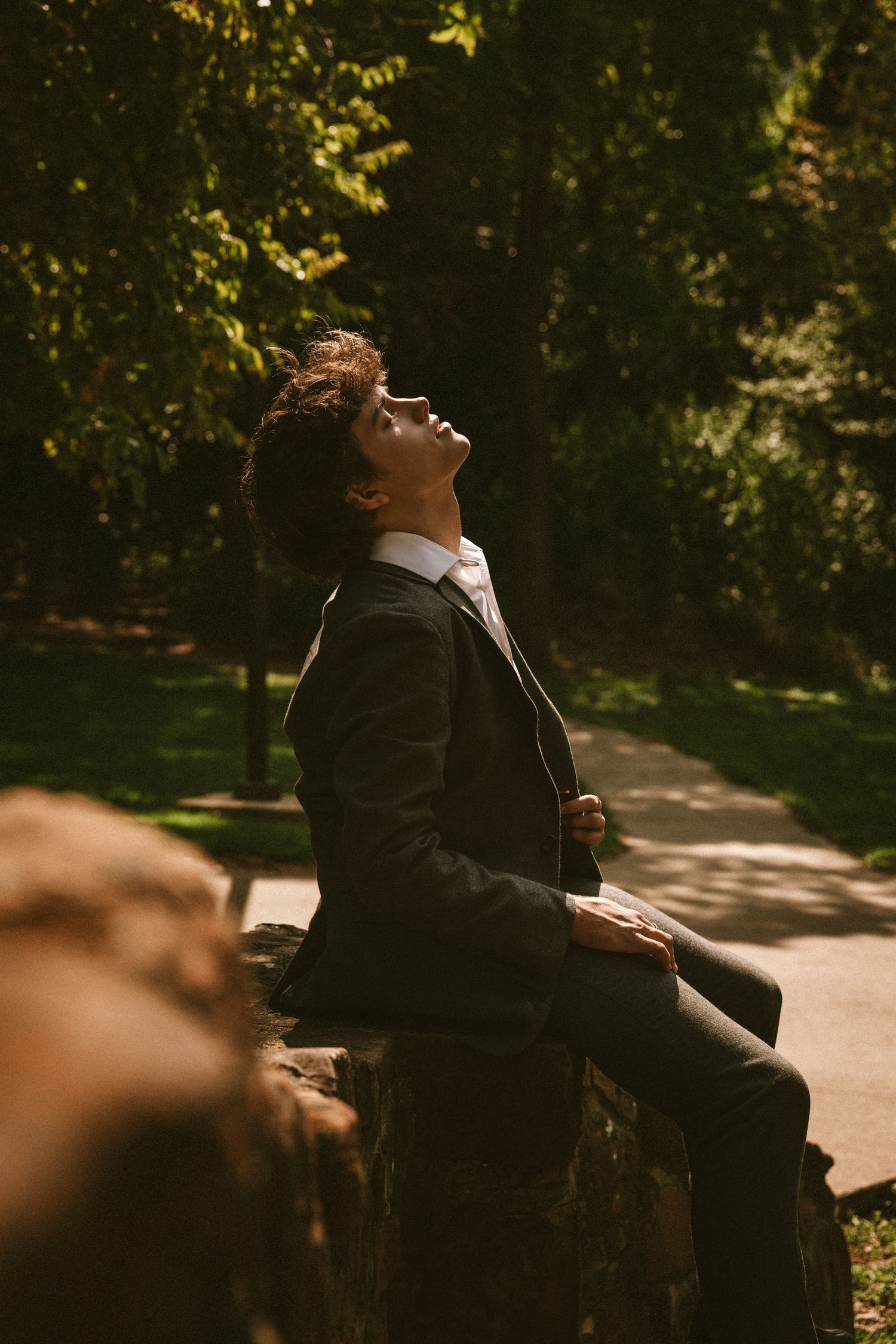 A person in a black jacket leans back on a bench in a sunlit park during autumn.