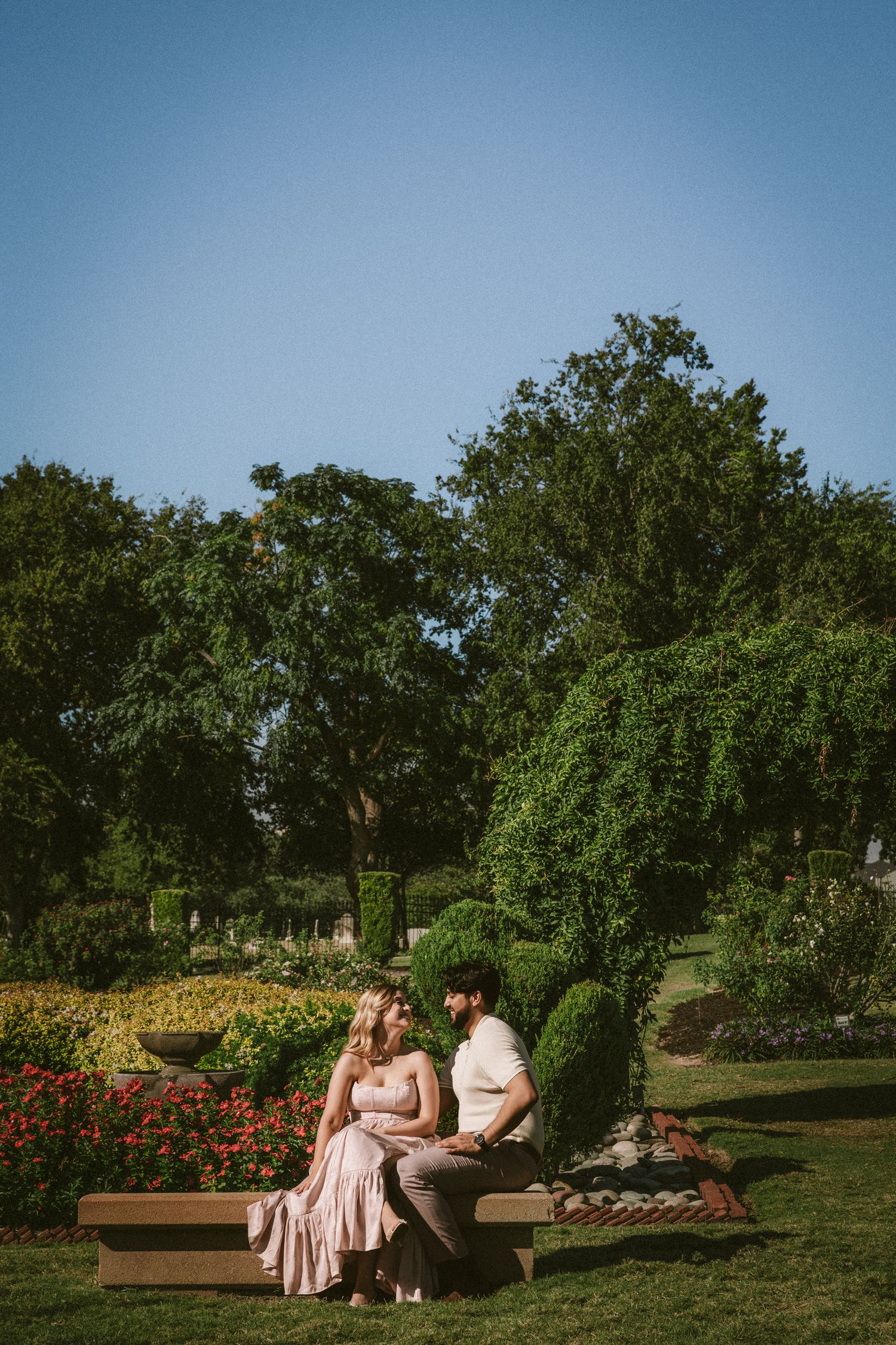 A romantic couple sits on a bench in a lush garden with blooming flowers and trees on a sunny afternoon.