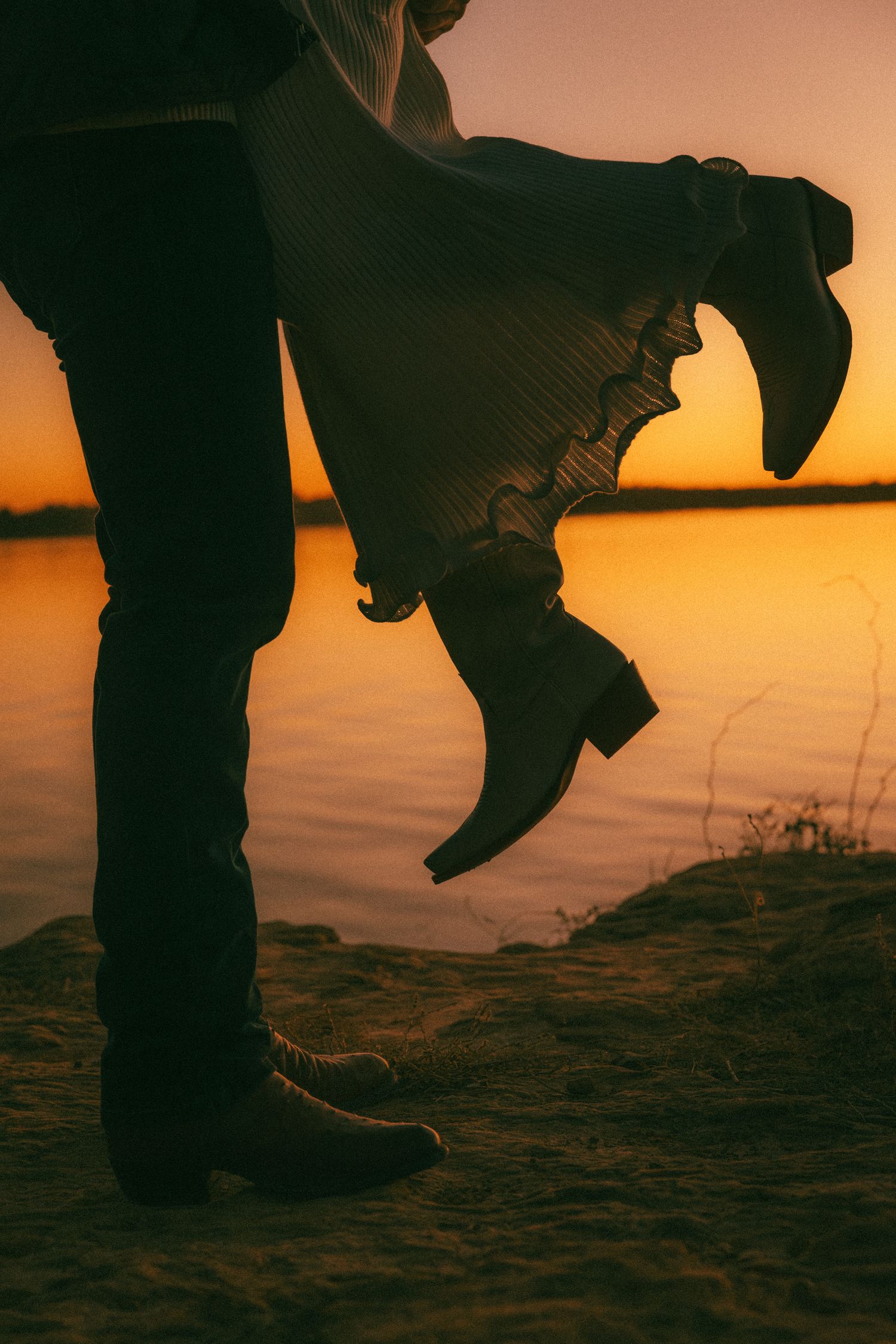 Silhouette of boots and flowing dress fabric against a golden sunset over water.