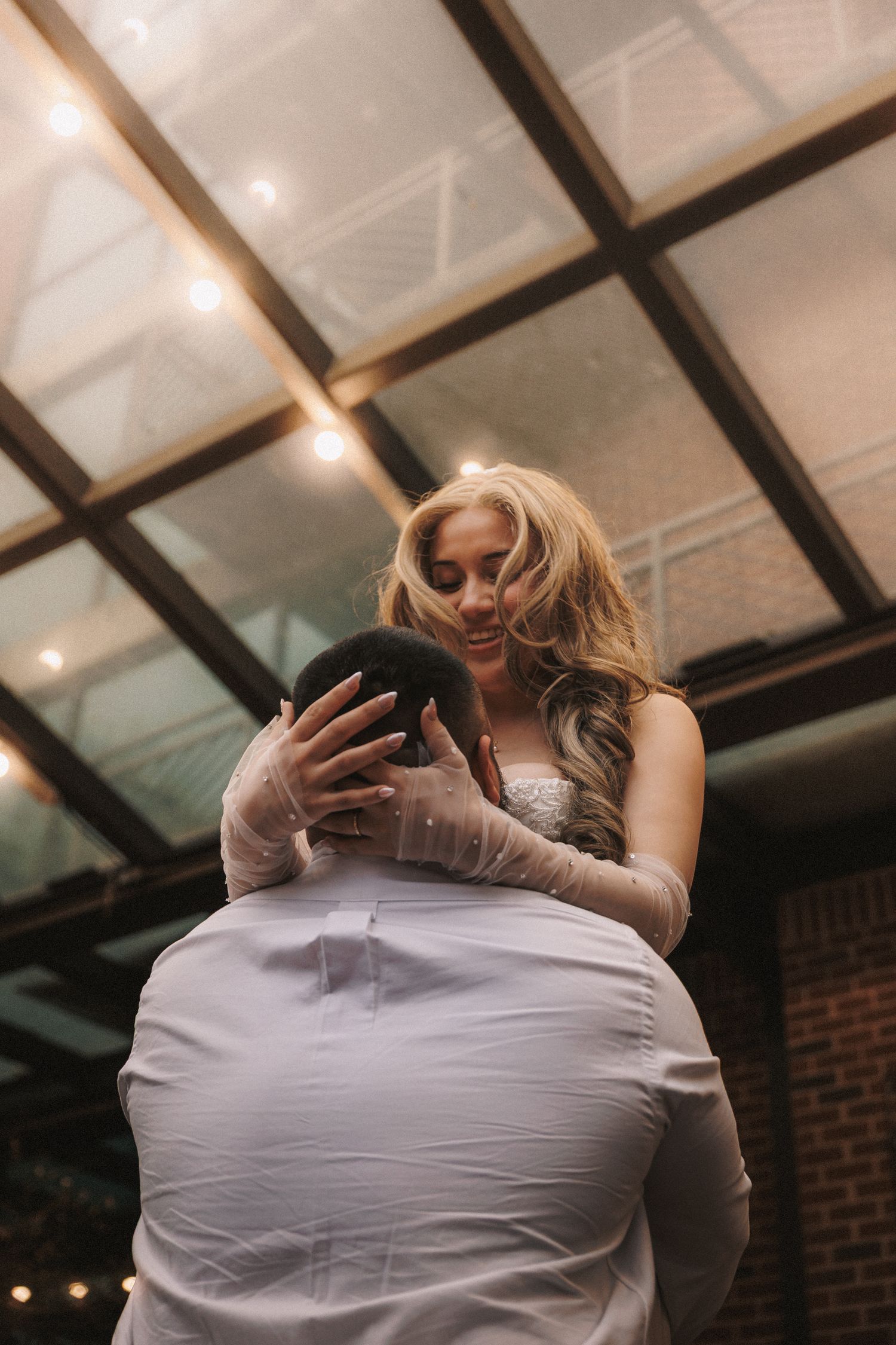 A romantic wedding dance moment captured under a glass ceiling with warm lighting and dramatic shadows.