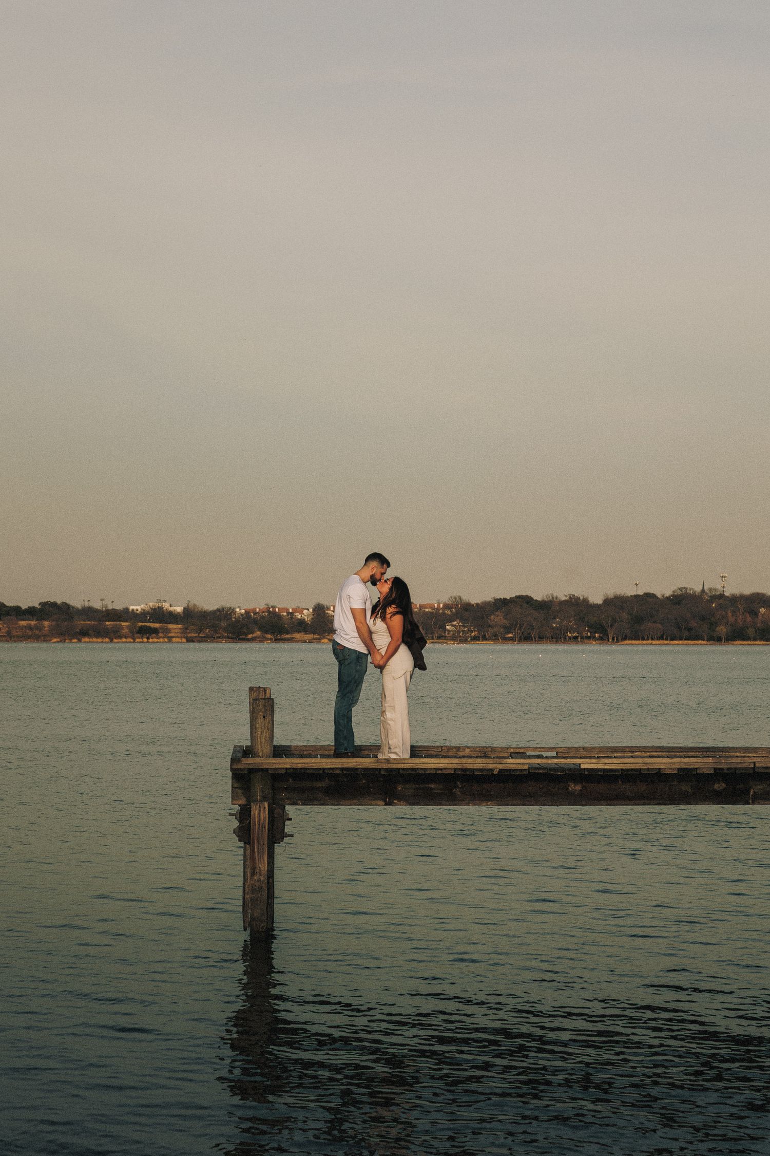 A couple stands on a wooden pier overlooking calm waters at sunset.