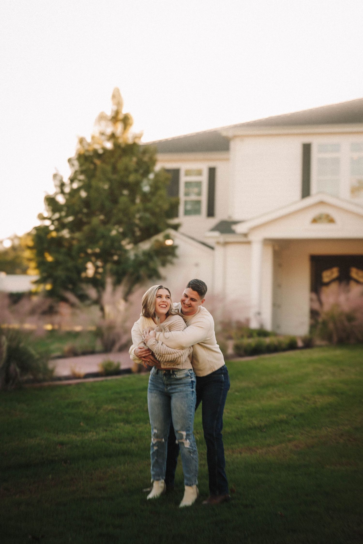 Loving couple laughs and embraces while standing together on a grassy lawn at sunset near suburban homes.