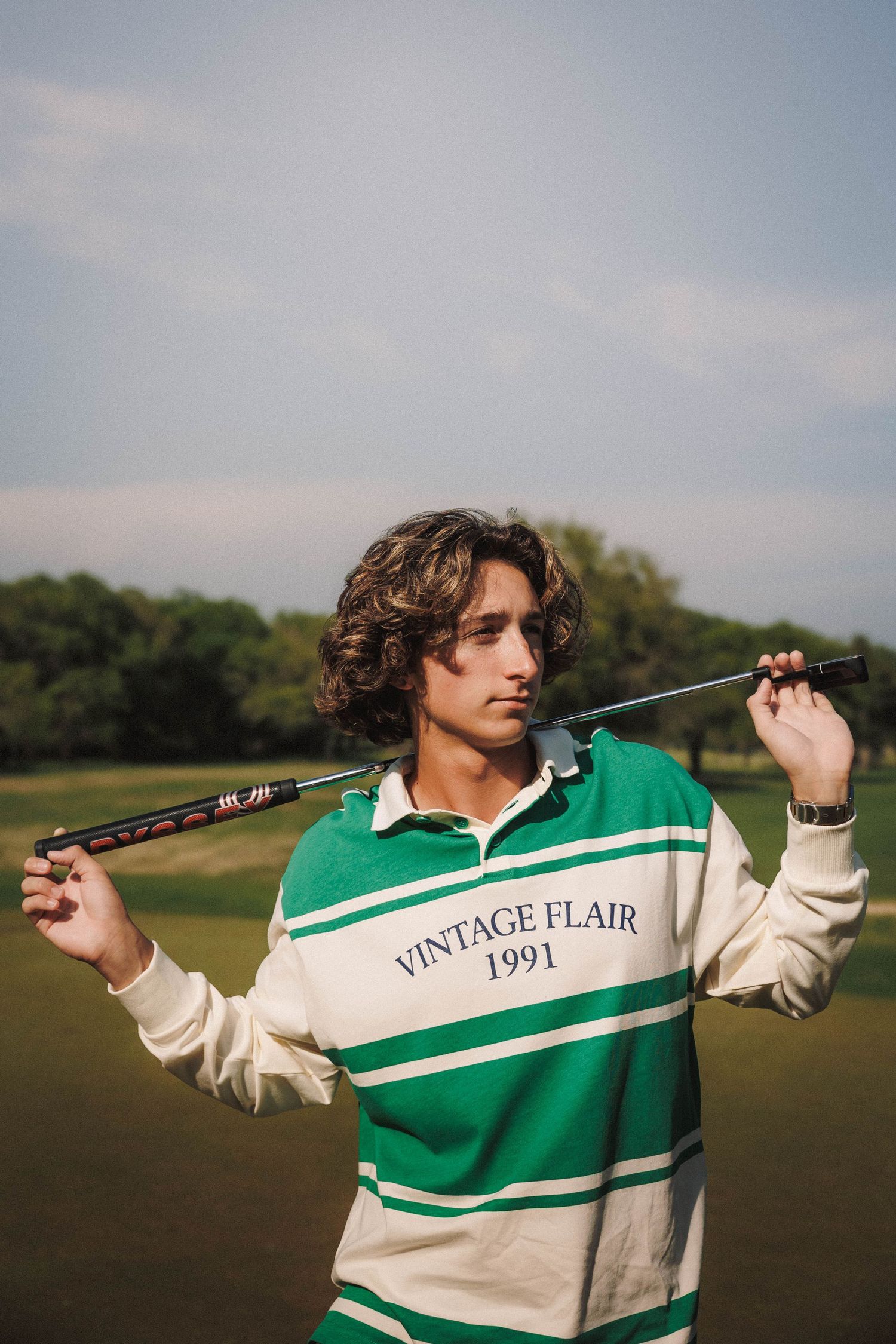 A person wearing a vintage-style green and white striped rugby shirt poses with a golf club on a grassy course.