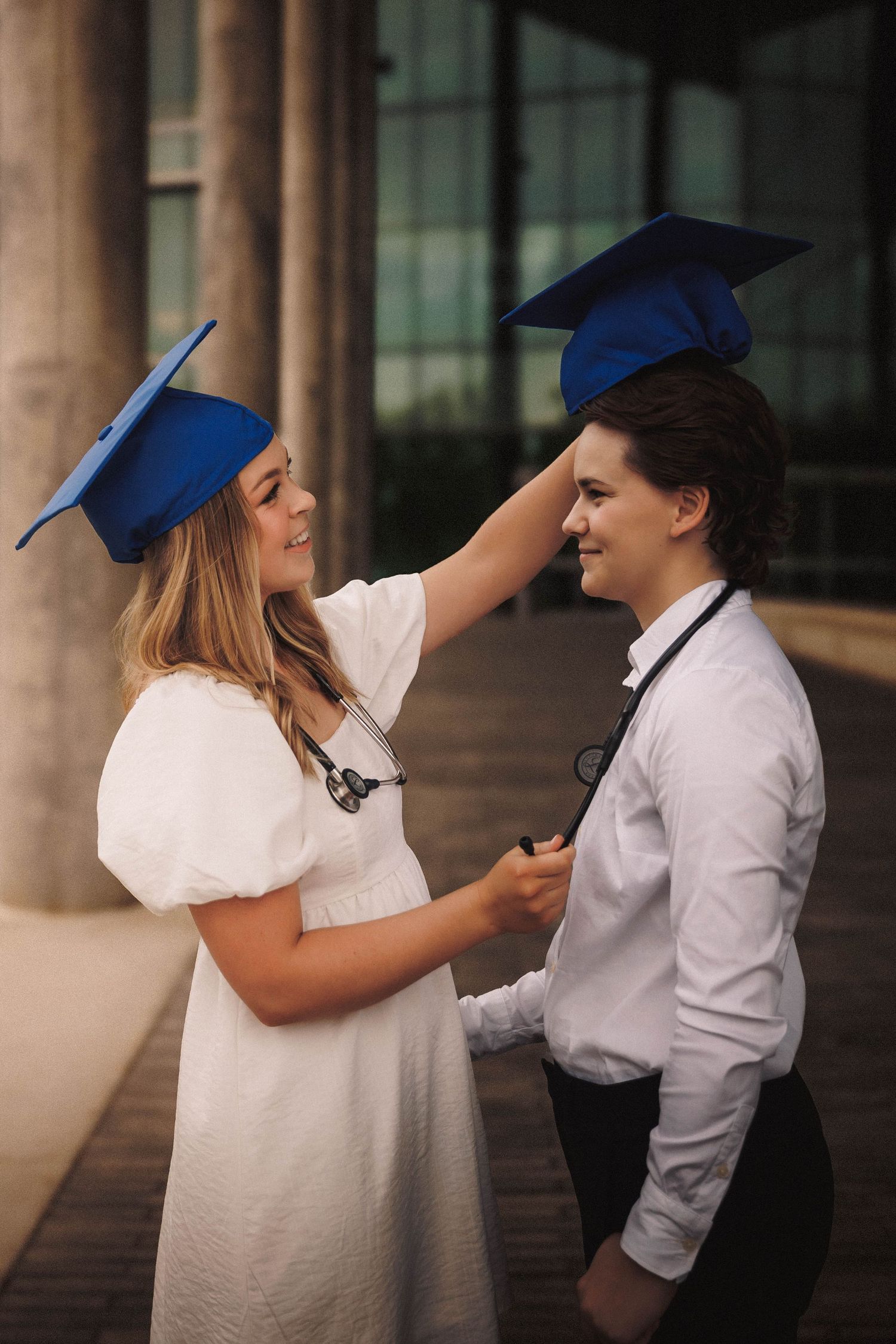 Two graduates in caps playfully interact outside building with pillared architecture.