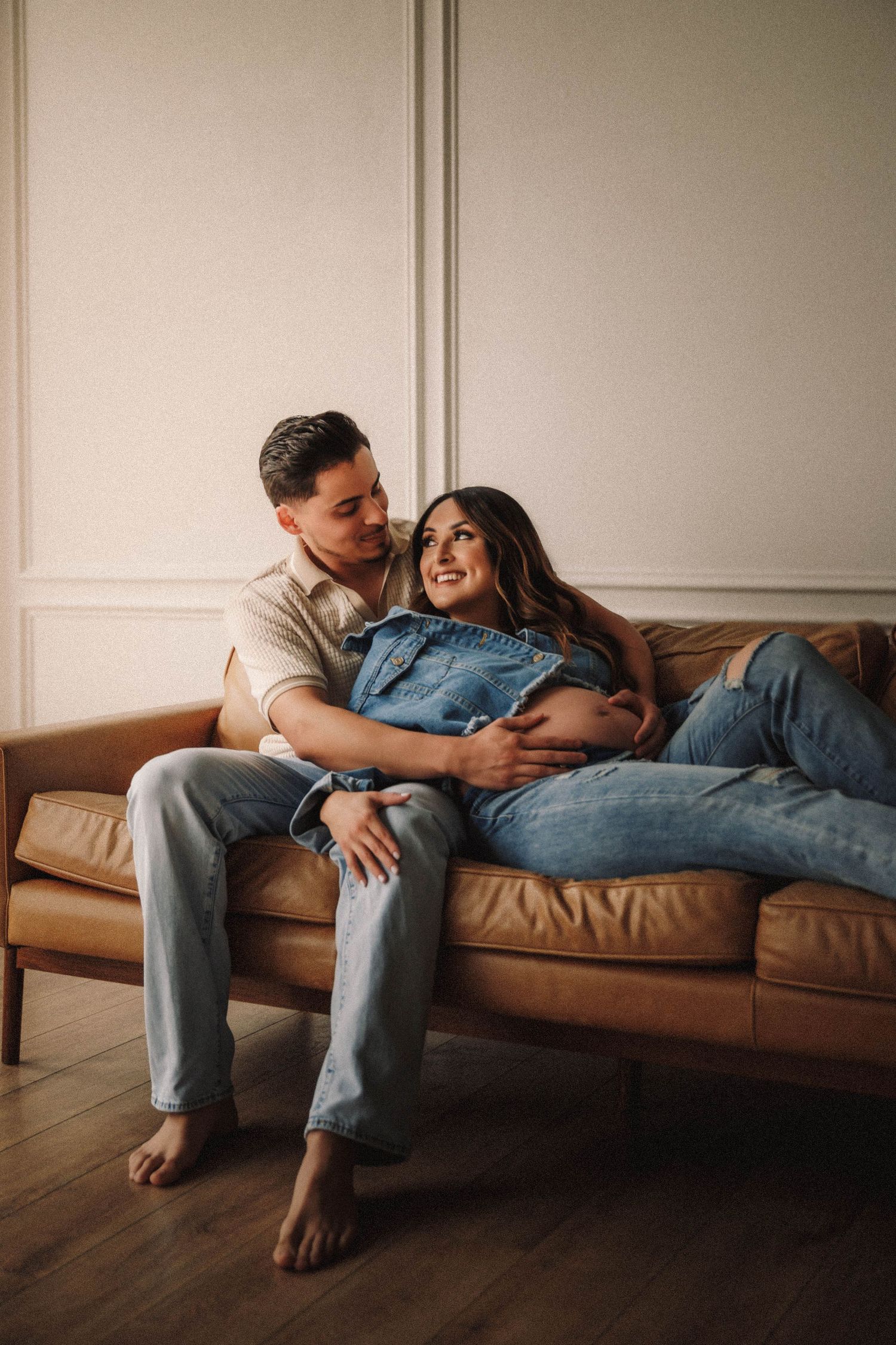 A couple shares an intimate moment while lounging together on a brown leather sofa in a minimalist room.