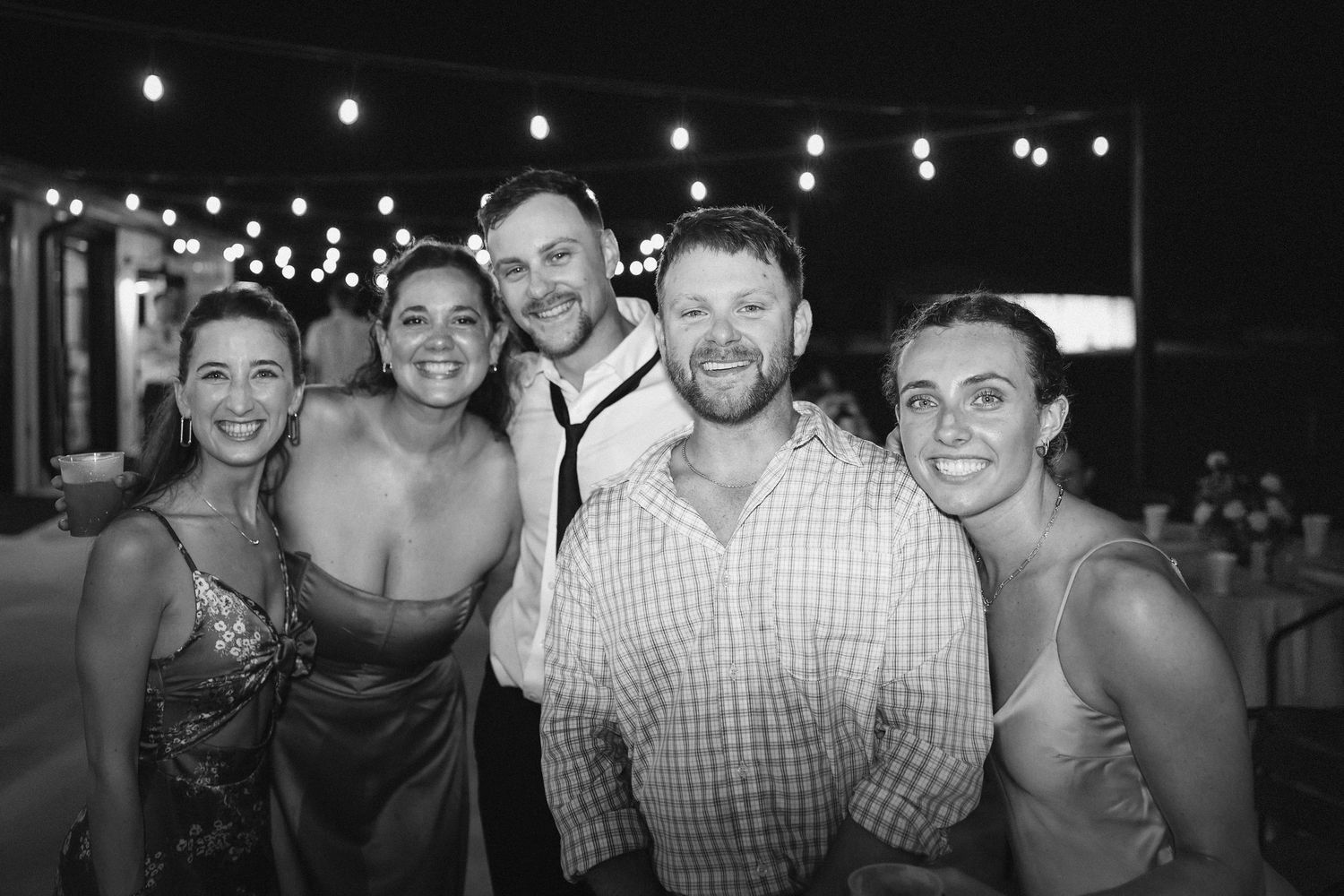 Wedding guests pose under string lights during evening reception celebration.