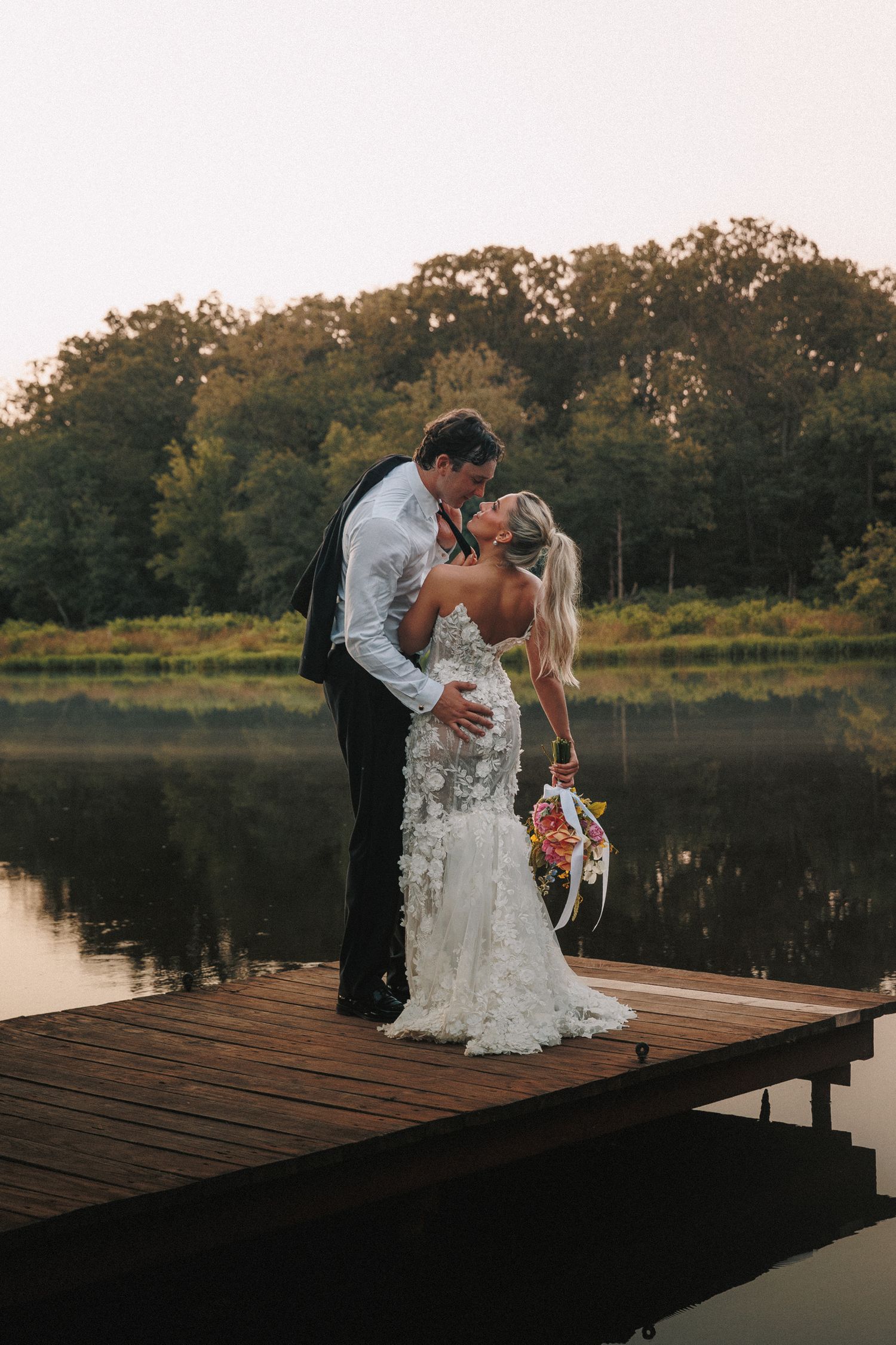 Newlyweds share an intimate moment on a wooden dock during sunset.