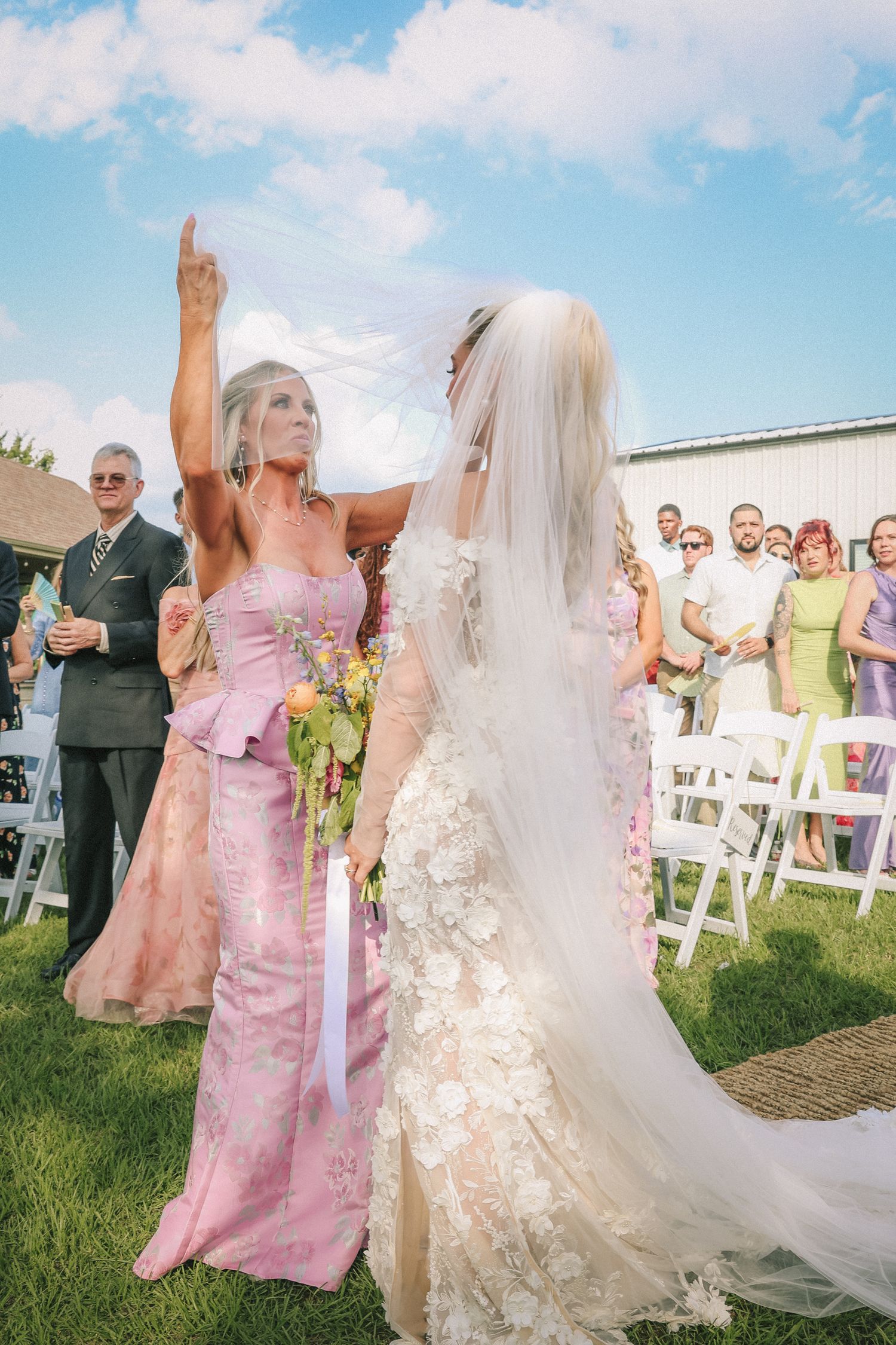 Wedding celebration with guests in pink dresses and white decor under blue sky.
