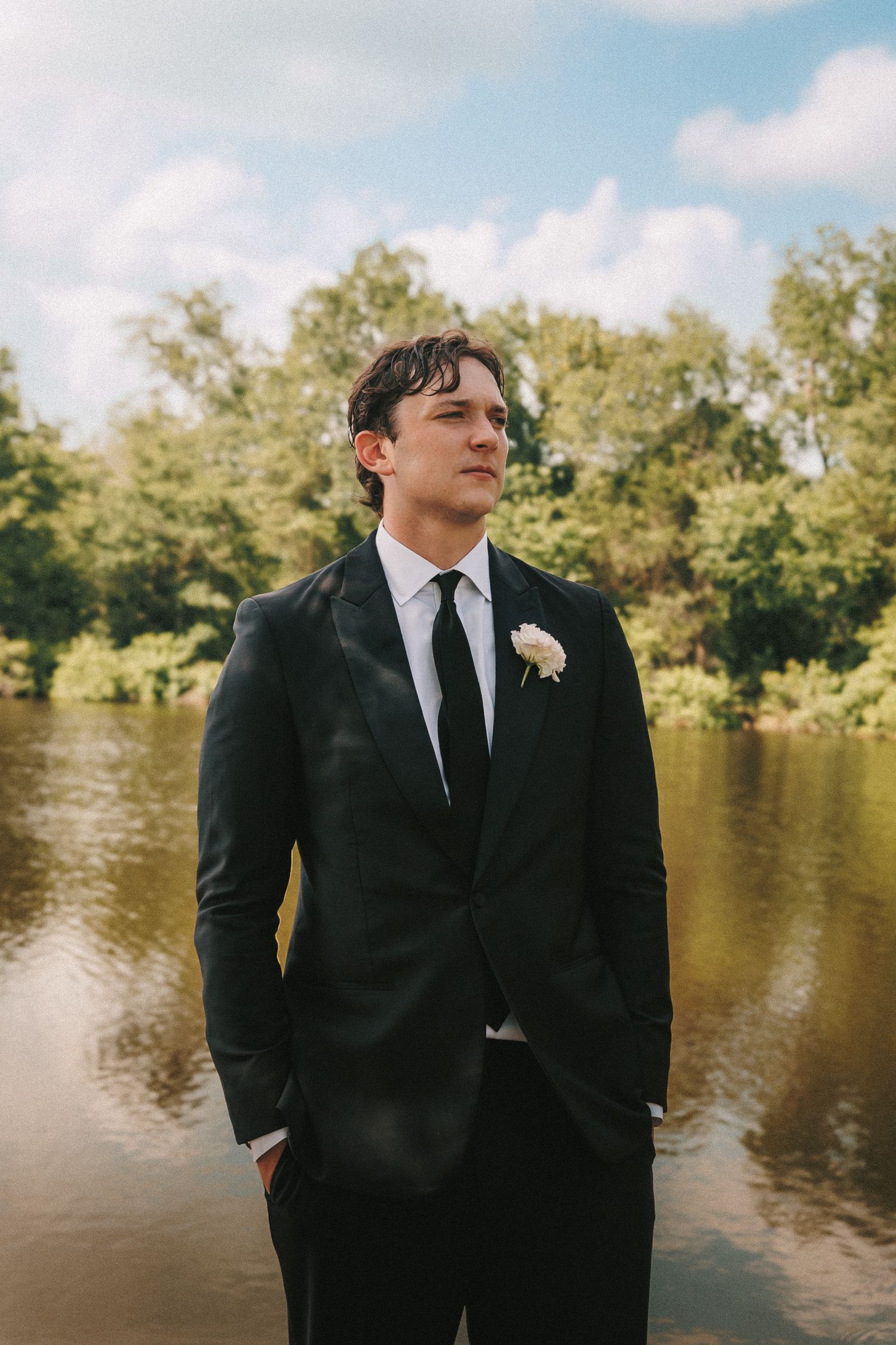 Groom in black suit poses near lake during wedding photoshoot.