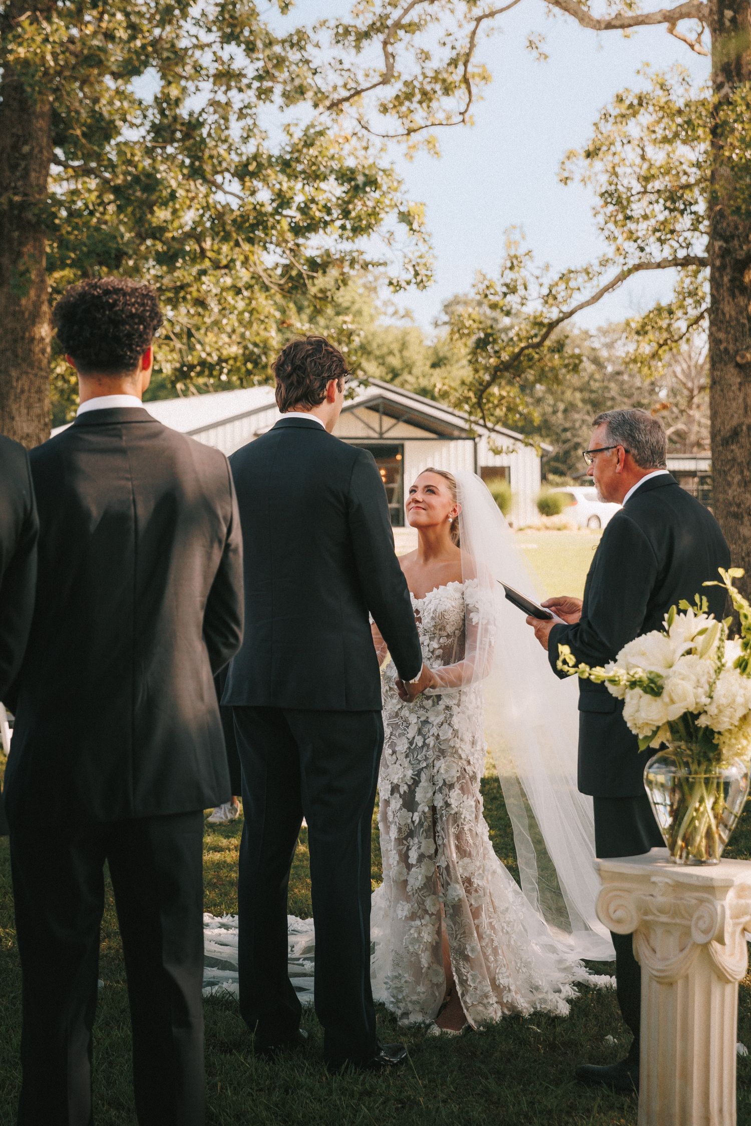 Wedding party members dressed in formal attire gather outdoors under trees during a ceremony.