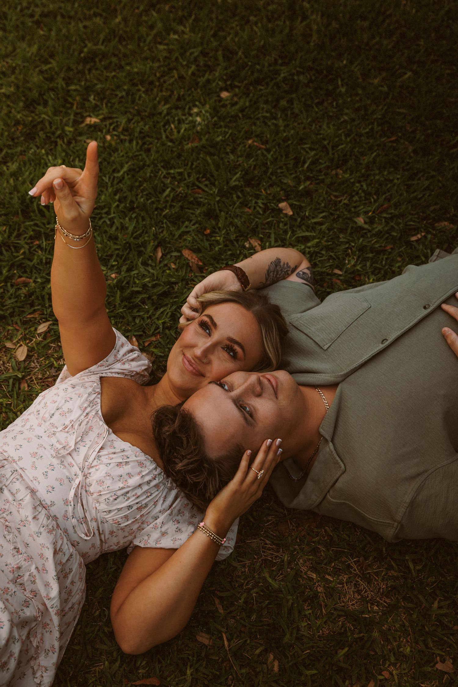 Two women lying on green grass, smiling and pointing up at the sky during a golden hour outdoor photo session.