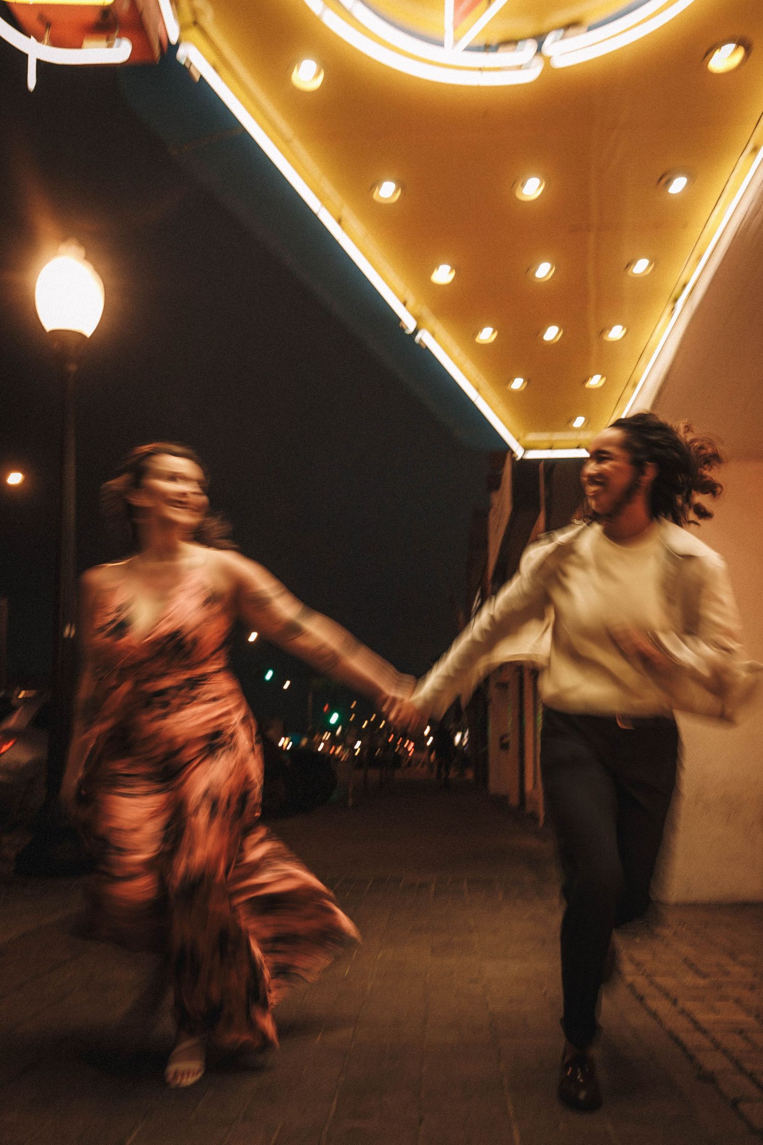 Newly engaged couple joyfully runs hand-in-hand under glowing theater marquee lights at night.