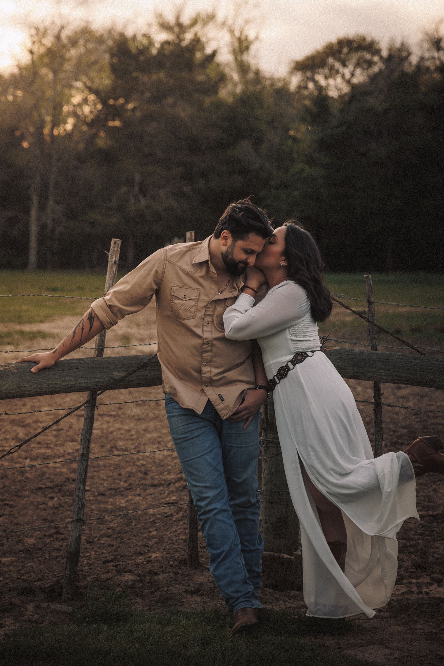 Couple sharing a tender moment leaning against a rustic wooden fence at sunset.