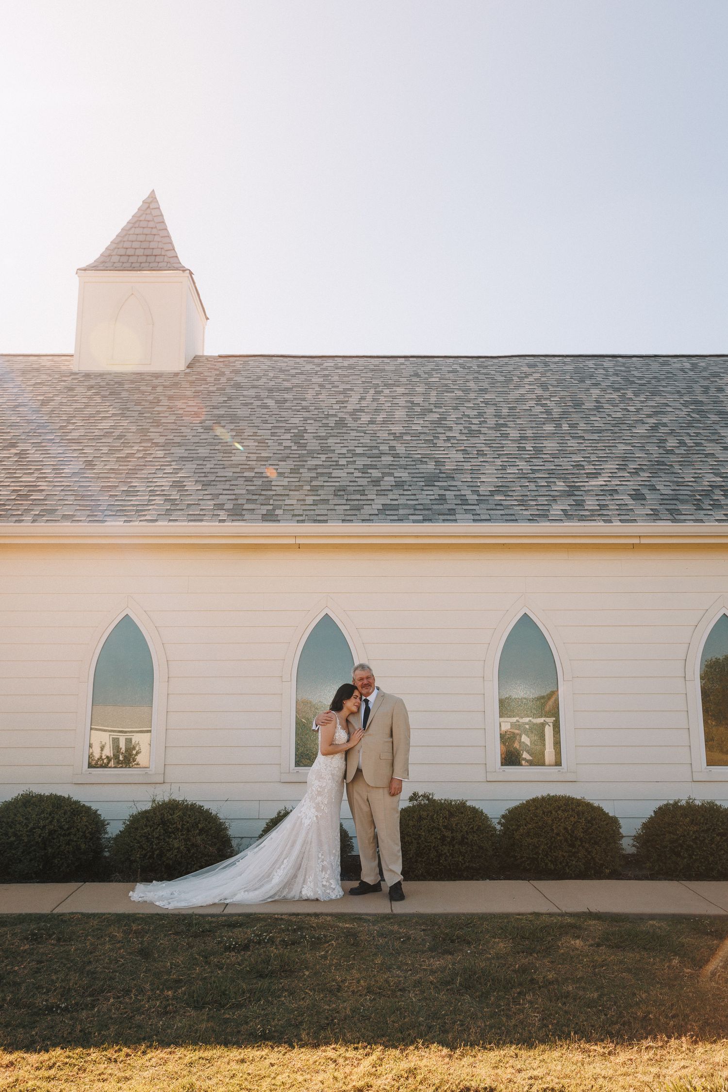 Bride and groom pose romantically outside a white chapel with gothic arched windows on their wedding day.