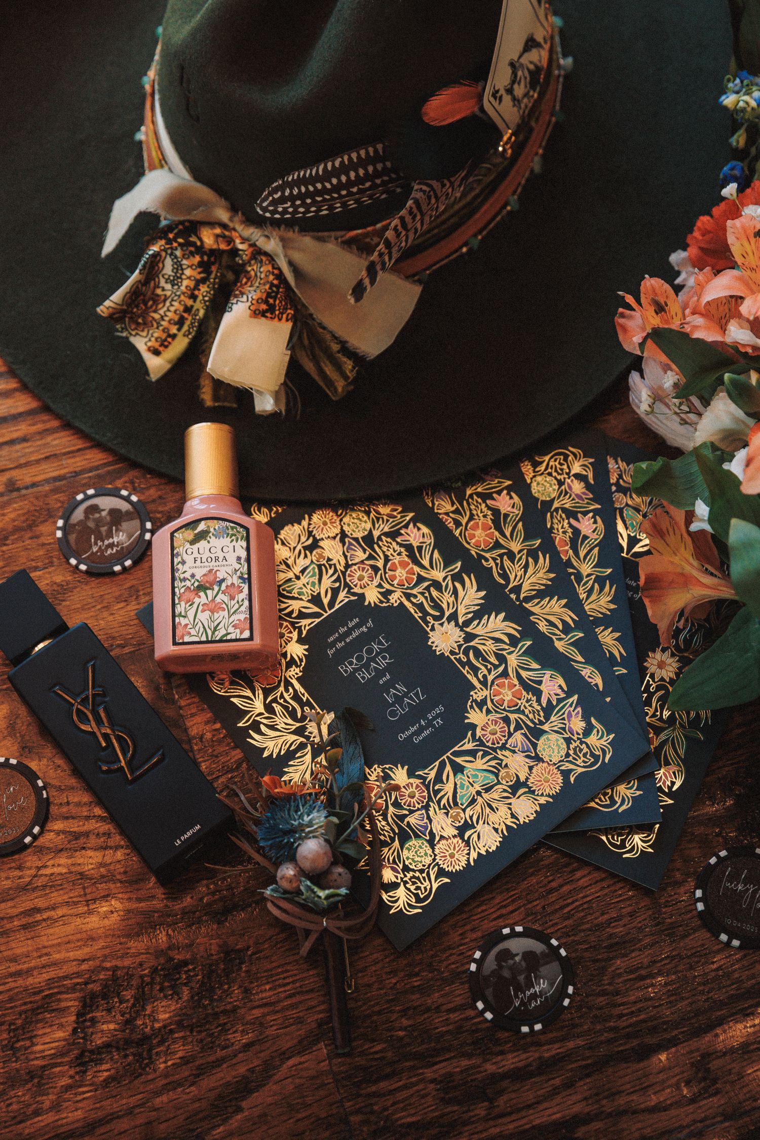 Boho wedding flat lay with floral invitations, Gucci perfume, YSL lipstick, and feathered hat.
