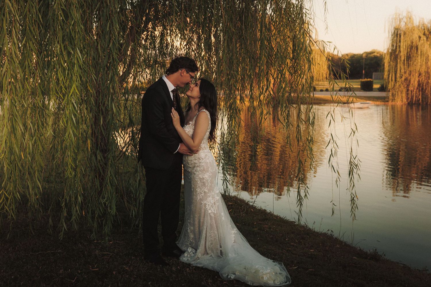 Bride and groom share an intimate moment under a weeping willow tree by a golden sunset lake.