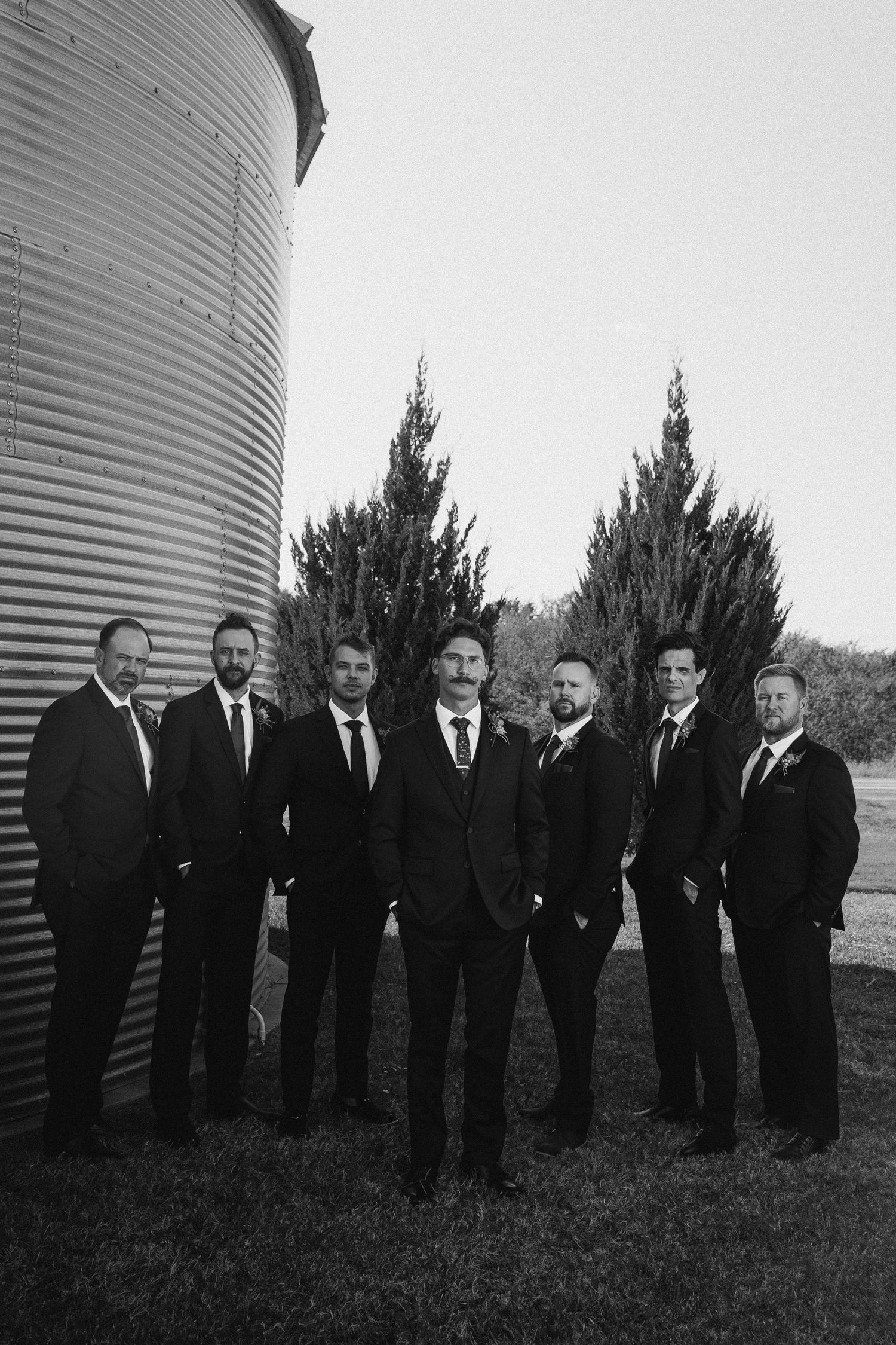 Black and white photo of groom and groomsmen in suits near a grain silo.