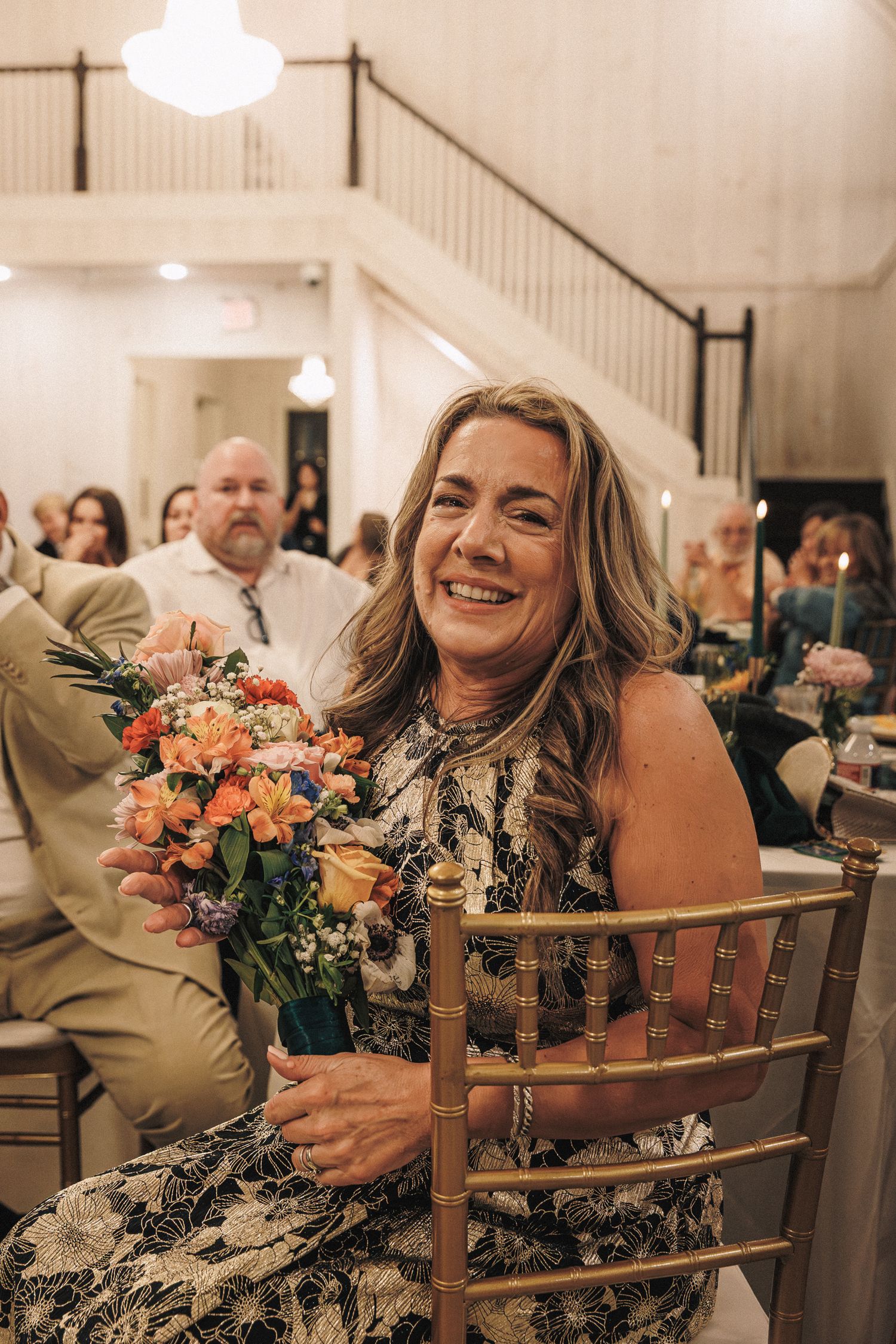 Smiling woman in floral dress holds a colorful bouquet of orange and pink flowers while seated at the wedding reception.