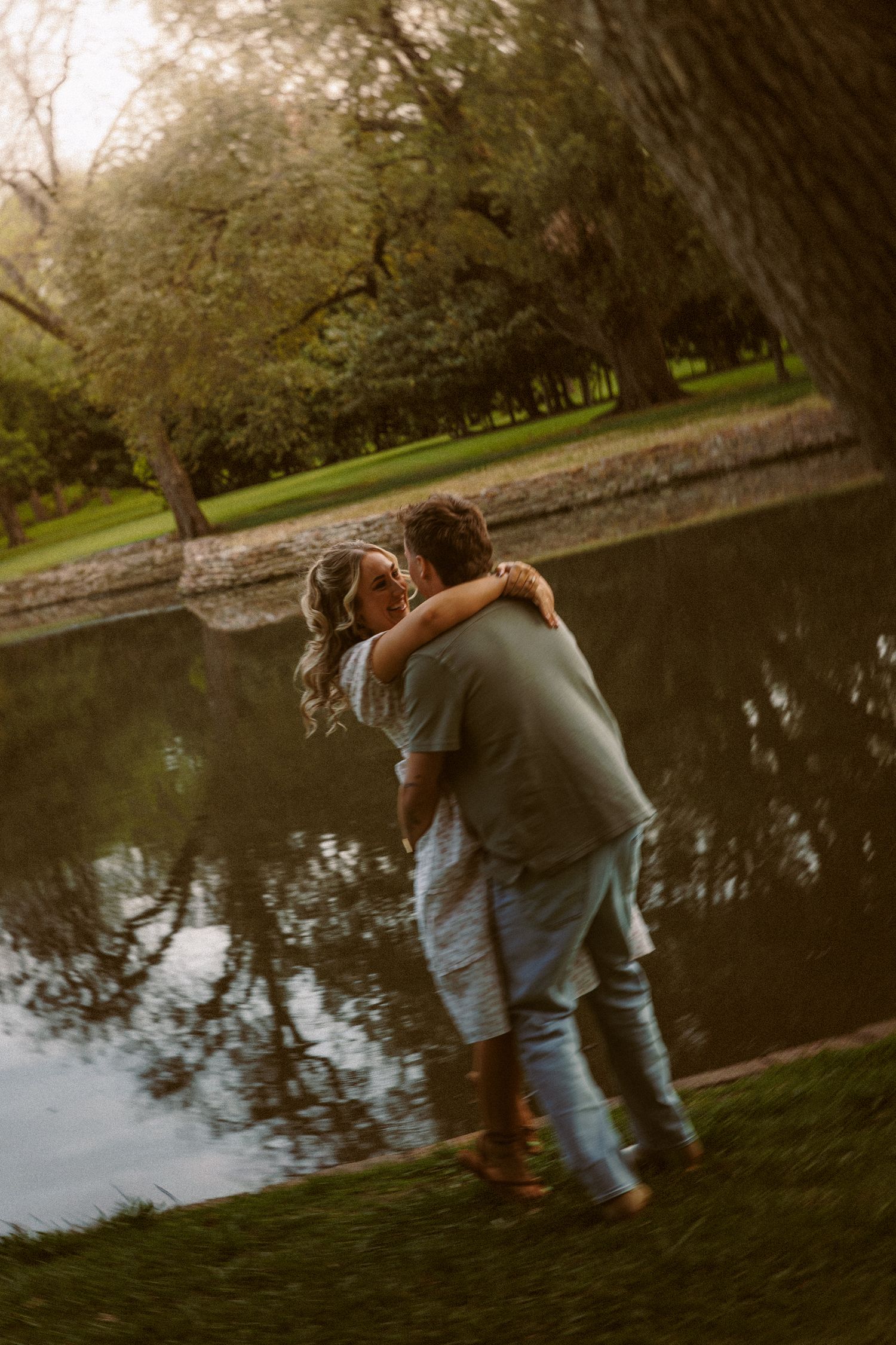 Romantic couple hugging and spinning near a reflective pond surrounded by trees at dusk.