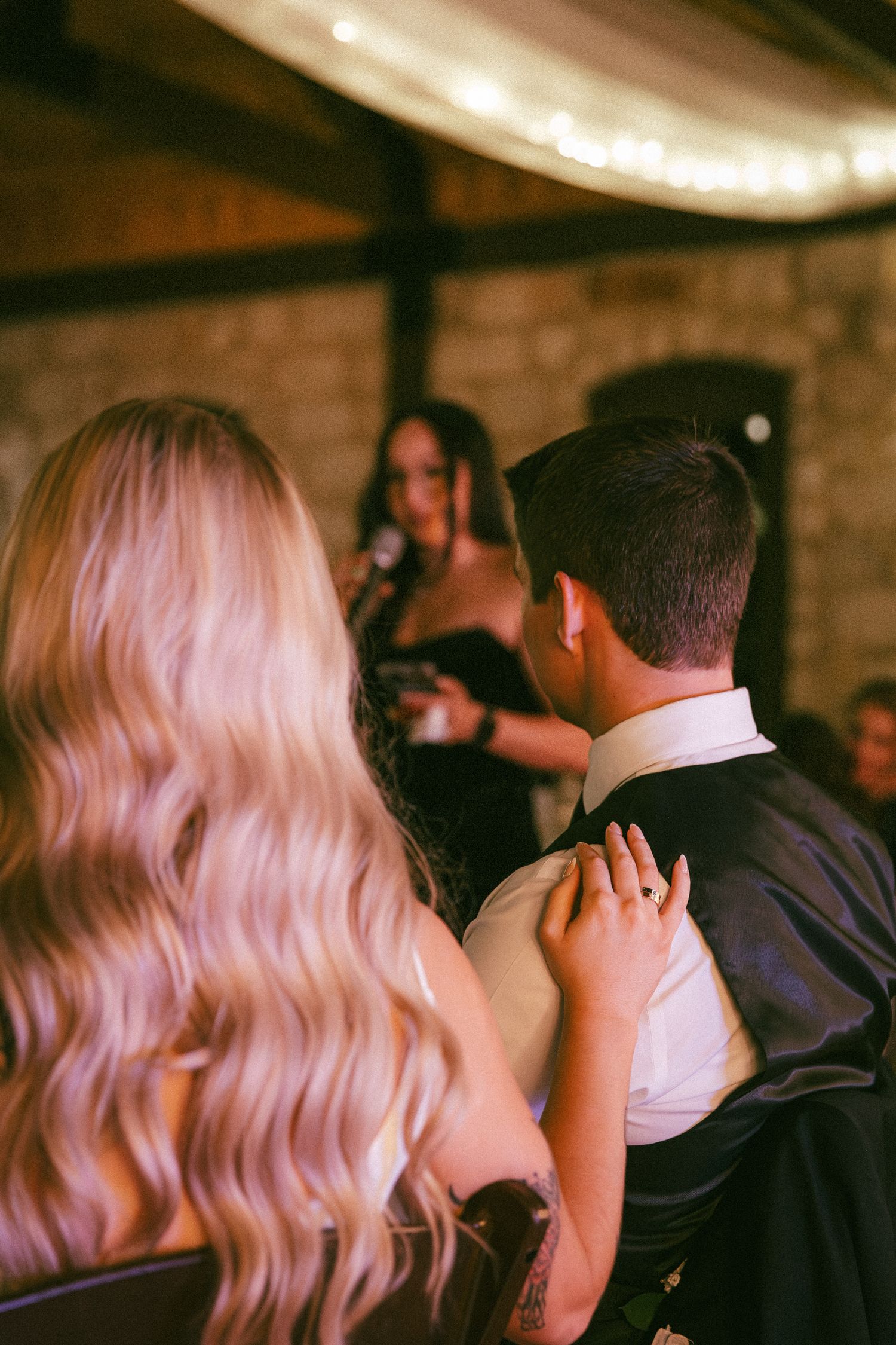 Bride rests hand on grooms shoulder while listening to speech at warmly lit wedding reception.