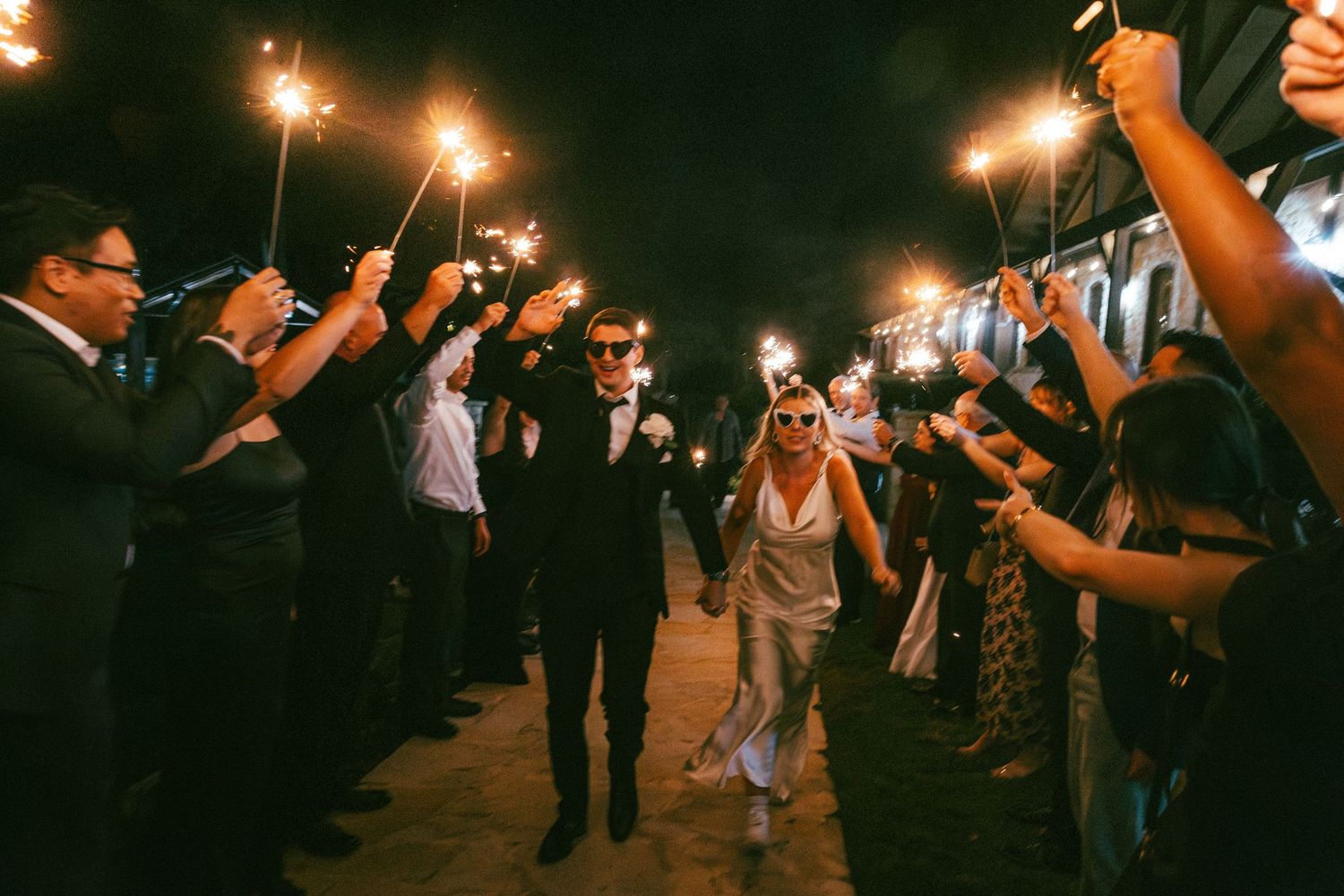 Newlyweds walking through sparkler send-off tunnel with cheering guests at night wedding exit.