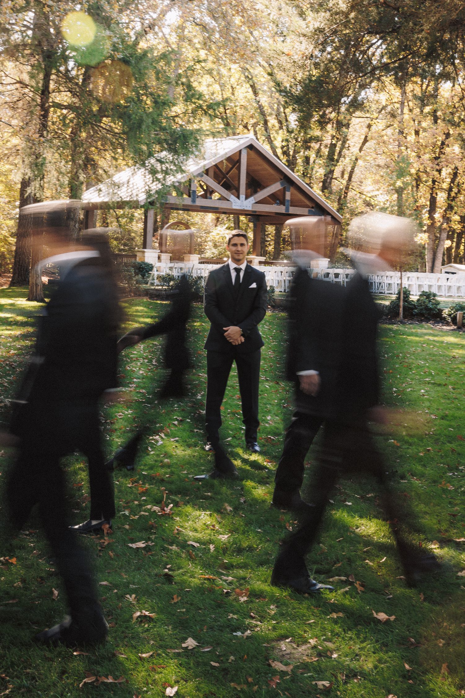 Groom stands still in black suit as groomsmen walk past in motion blur outdoors at autumn wedding venue.