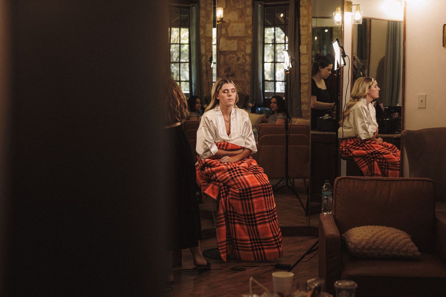 Woman in plaid skirt and white blouse sits pensively in a warmly lit rustic interior with mirrors.