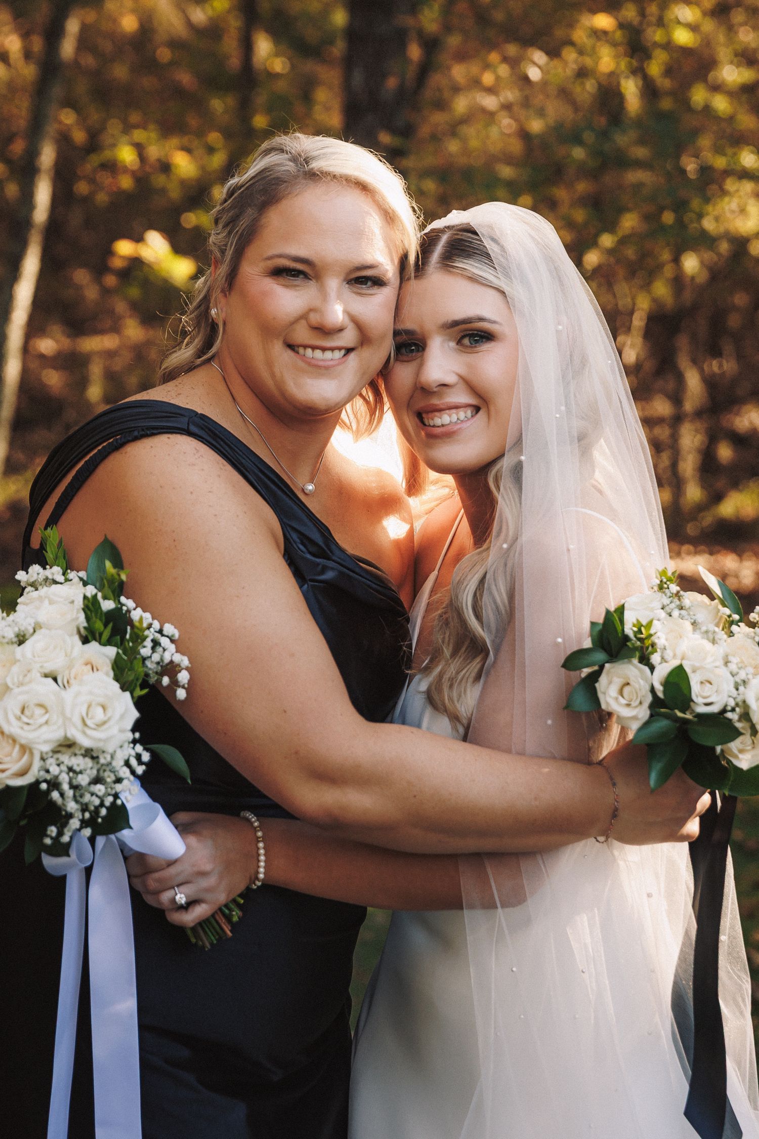 Smiling bride in white gown and veil embracing bridesmaid in navy dress holding white rose bouquets.