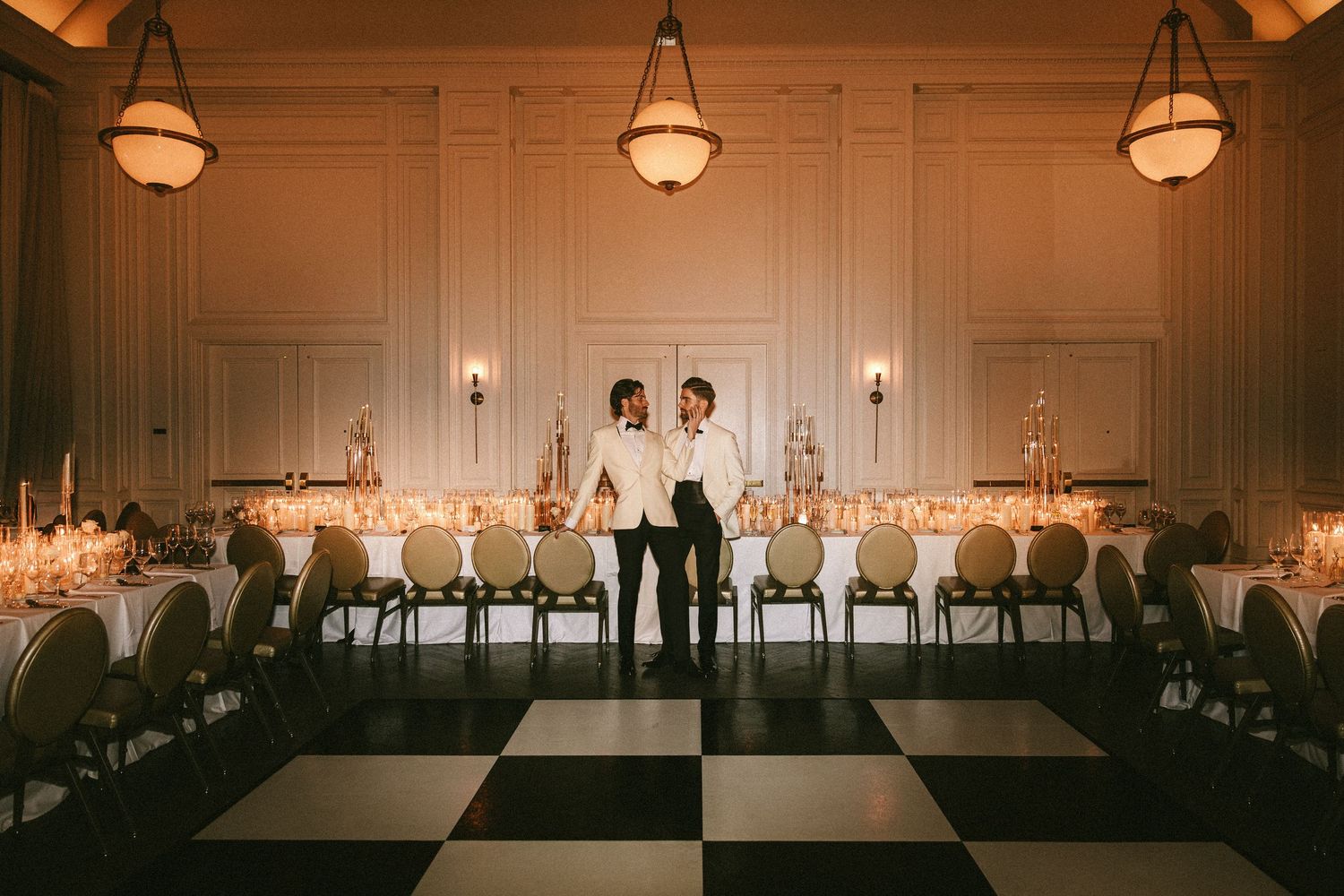 Two grooms stand on a black and white checkered dance floor in a grand ballroom.