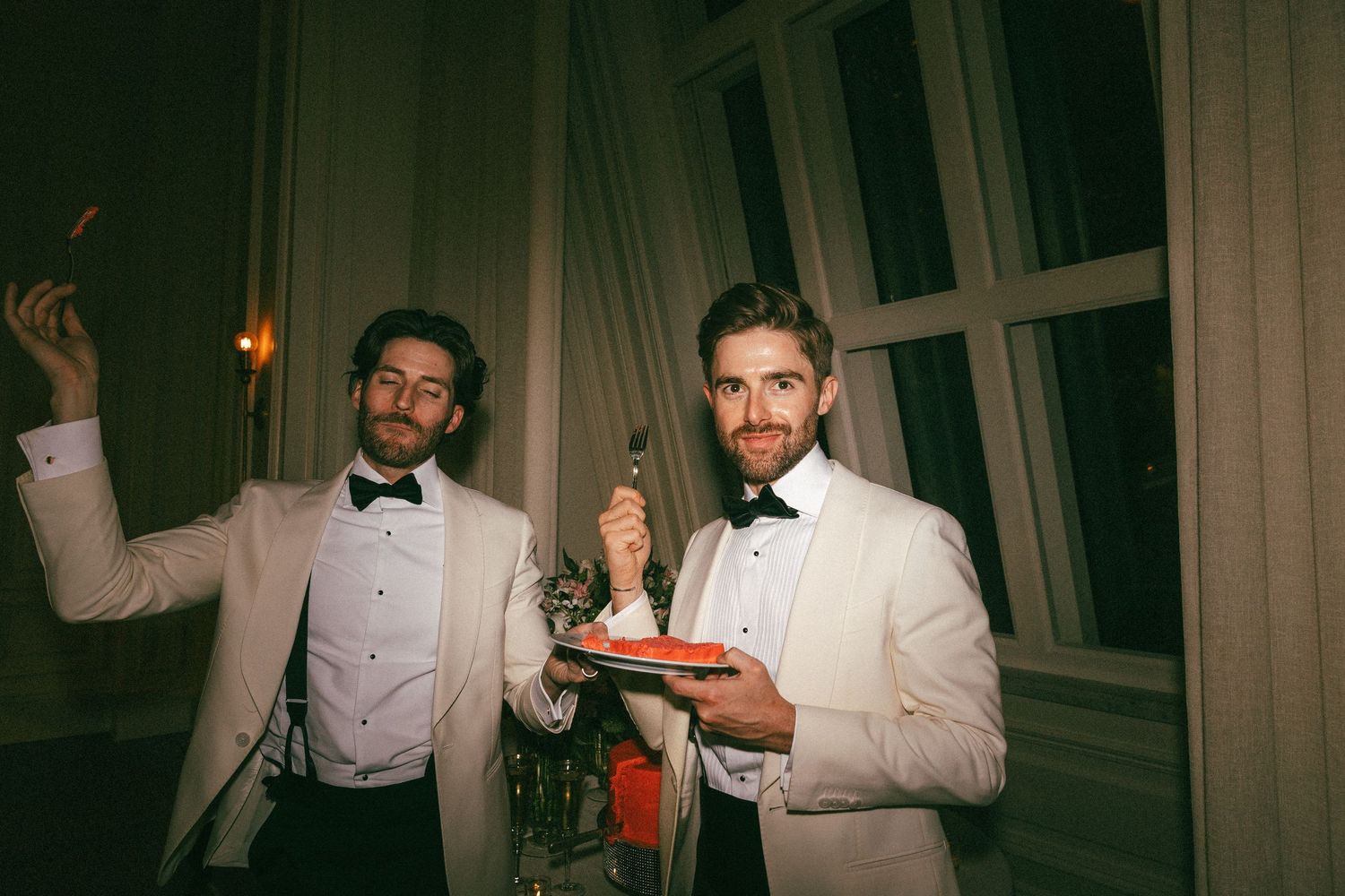 Two grooms in white tuxedo jackets smile and celebrate at their glamorous evening wedding reception.